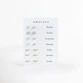 Set Daily Edit de Amalaya compuesto por siete pares de aretes enchapados en oro 14k que incluyen cuatro pares de broqueles de circonia y tres pares de arracadas huggies, vista frontal sobre fondo blanco, Vista de frente del juego semanal de 7 pares de aretes en tono oro de 14k, con broqueles solitarios de 0.3 cm a 0.6 cm y arracadas minimalistas de 0.9 cm a 1 cm, Plano general de set de aretes dorados semanales presentados en tarjeta blanca con los días de la semana
