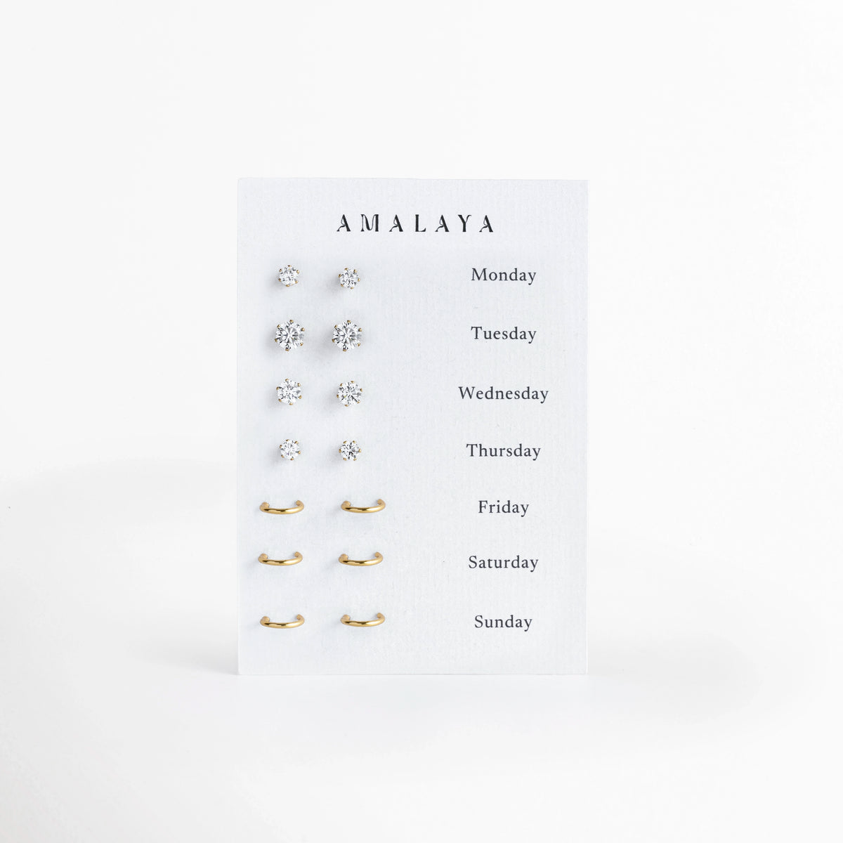 Set Daily Edit de Amalaya compuesto por siete pares de aretes enchapados en oro 14k que incluyen cuatro pares de broqueles de circonia y tres pares de arracadas huggies, vista frontal sobre fondo blanco, Vista de frente del juego semanal de 7 pares de aretes en tono oro de 14k, con broqueles solitarios de 0.3 cm a 0.6 cm y arracadas minimalistas de 0.9 cm a 1 cm, Plano general de set de aretes dorados semanales presentados en tarjeta blanca con los días de la semana