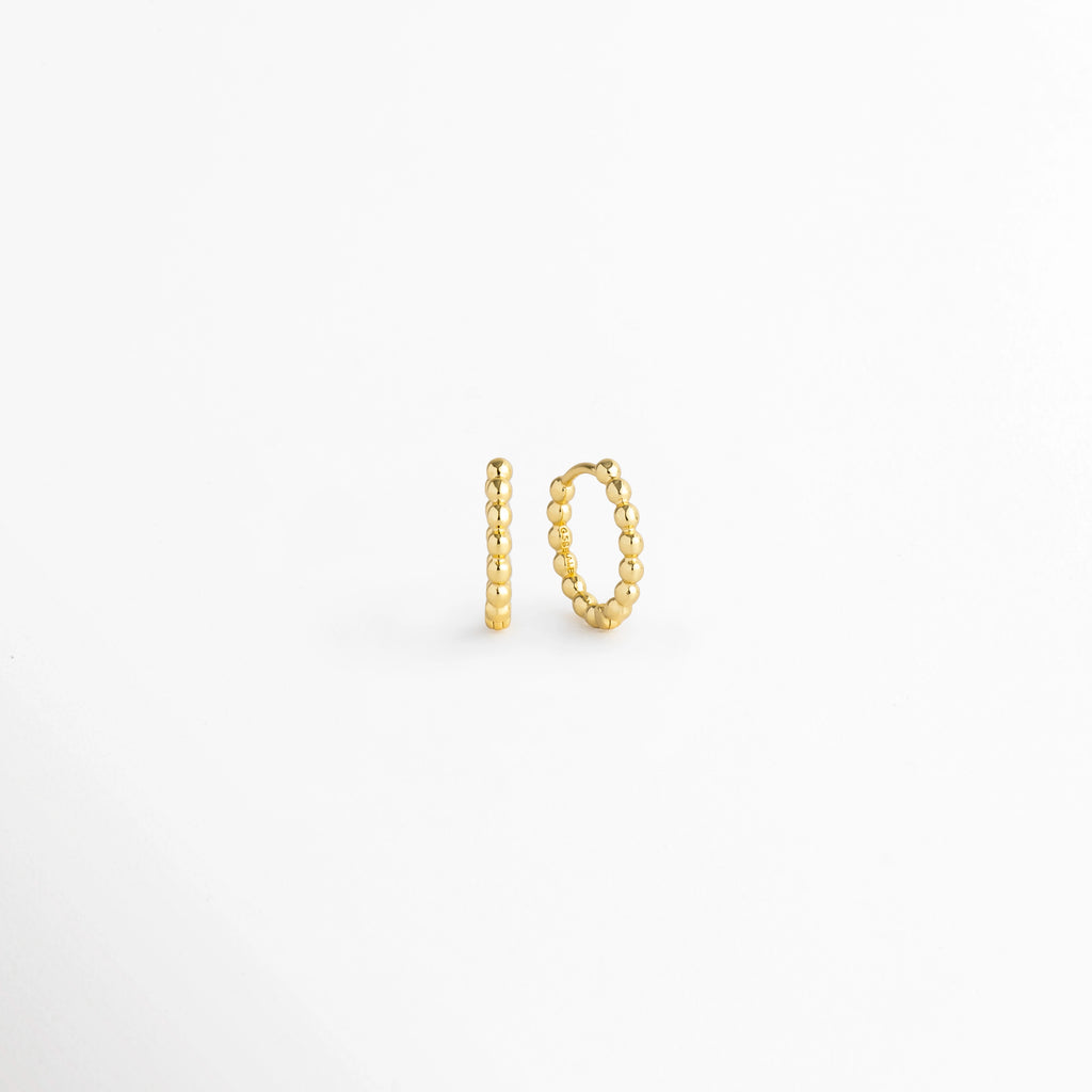 Arracadas Golden Hoops vistas de perfil y tres cuartos mostrando la silueta circular perlada y la Bisagra de apertura, Vista en ángulo de aretes tipo huggie dorados resaltando la textura de cuentas redondeadas de su superficie, Acercamiento a la silueta circular con textura de bolitas doradas en toma de tres cuartos sobre fondo claro, Detalle del diámetro compacto de la arracada perlada Golden Hoops enchapada en oro.