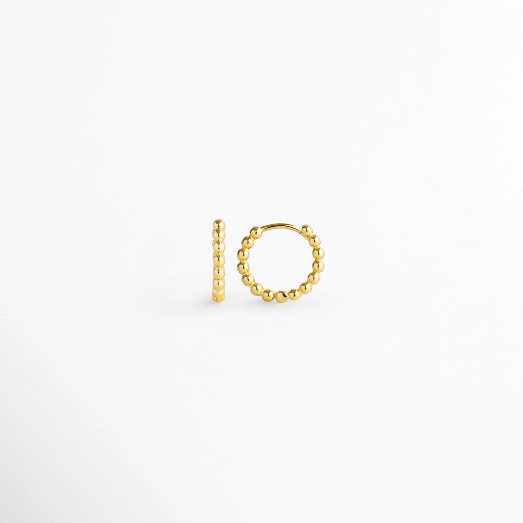 Vista lateral de la arracada Golden Hoops con cierre de bisagra/huggie encajado firmemente en el poste, Detalle del mecanismo de cierre click/snap oculto en arracada con textura de bolitas, Muestra lateral del perfil arqueado y bordes perlados de la arracada dorada sobre superficie blanca, Vista de cerca del poste delgado y la curvatura del aro pequeño con relieve de cuentas.