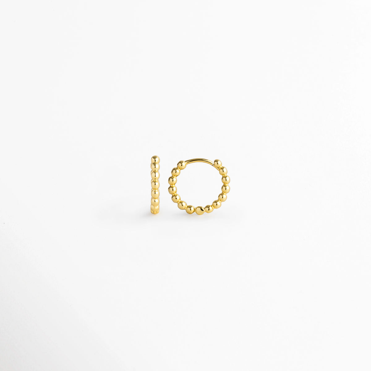 Vista lateral de la arracada Golden Hoops con cierre de bisagra/huggie encajado firmemente en el poste, Detalle del mecanismo de cierre click/snap oculto en arracada con textura de bolitas, Muestra lateral del perfil arqueado y bordes perlados de la arracada dorada sobre superficie blanca, Vista de cerca del poste delgado y la curvatura del aro pequeño con relieve de cuentas.