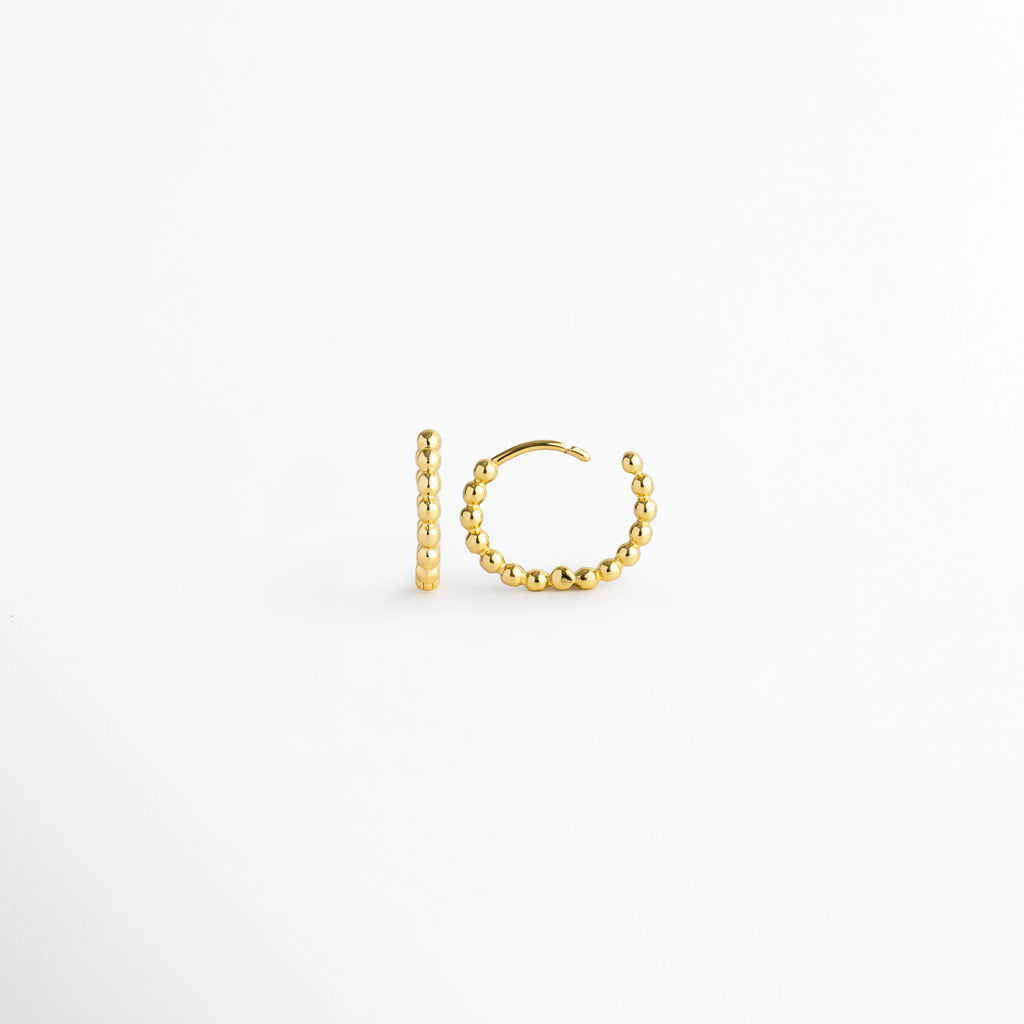 Vista lateral de la arracada Golden Hoops con la bisagra completamente abierta mostrando el poste de inserción, Acercamiento al broche articulado abierto de la arracada dorada con textura de bolitas continuas, Detalle del sistema de apertura fácil de la arracada estilo huggie sobre fondo claro.