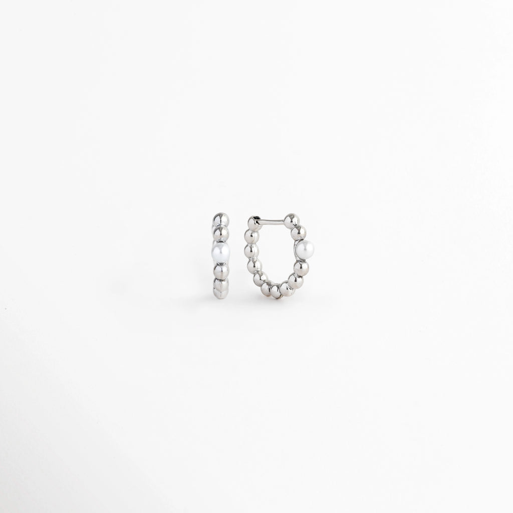 Vista en perspectiva de los Silver Pearl Hoops mostrando la curvatura del aro Huggie y su mecanismo de cierre articulado, Acercamiento al broche de bisagra o Huggie del arete plateado en posición entreabierta sobre fondo blanco, Toma angular que resalta el grosor y el relieve esférico de las arracadas plateadas con perla, Muestra del perfil redondeado y la integración de la perla en la caña metálica electrochapada.