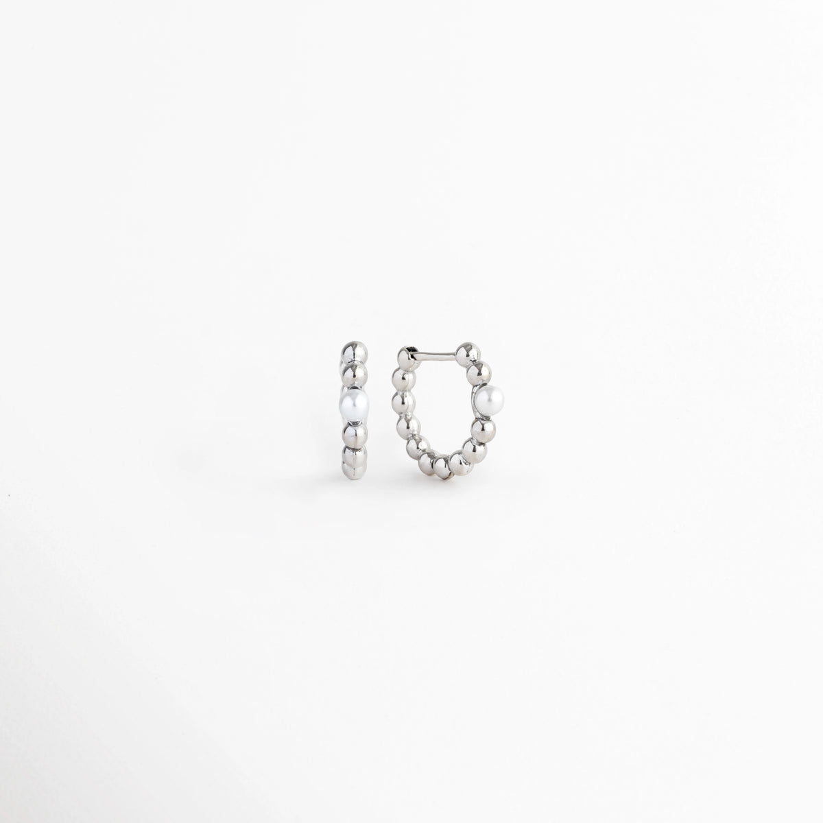 Vista en perspectiva de los Silver Pearl Hoops mostrando la curvatura del aro Huggie y su mecanismo de cierre articulado, Acercamiento al broche de bisagra o Huggie del arete plateado en posición entreabierta sobre fondo blanco, Toma angular que resalta el grosor y el relieve esférico de las arracadas plateadas con perla, Muestra del perfil redondeado y la integración de la perla en la caña metálica electrochapada.