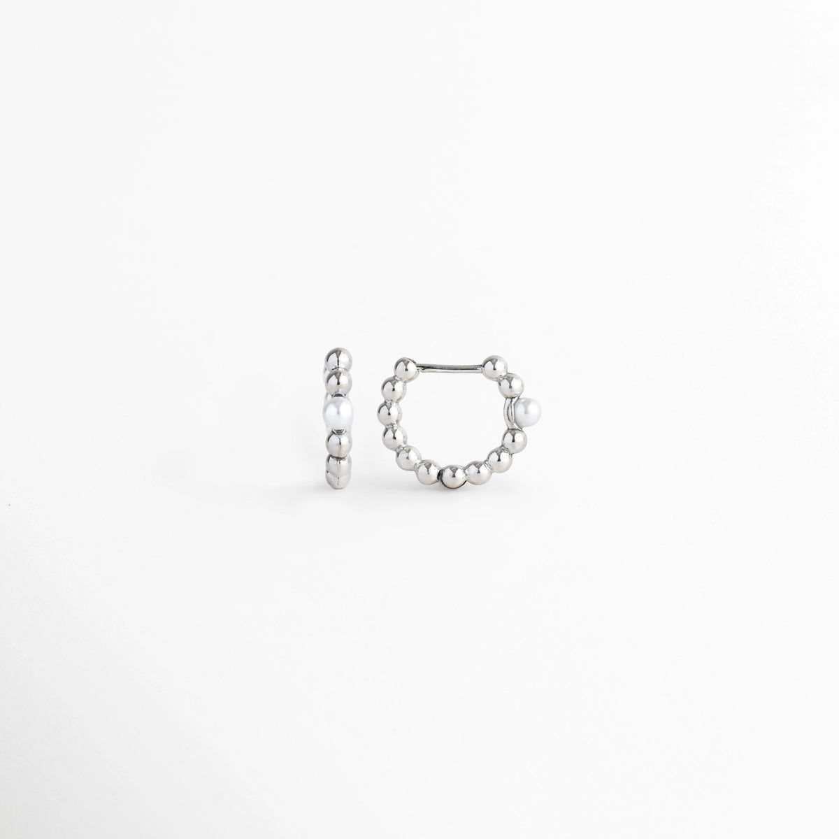 Vista lateral de los Silver Pearl Hoops exhibiendo el perfil circular compacto y el mecanismo de cierre a presión, Acercamiento de lado a la curvatura del aro metálico plateado con cuentas alineadas, Toma lateral que muestra el poste delgado articulado para un ajuste seguro en la oreja, Muestra del acabado electrochapado en tono plata pulido y la simetría del aro.