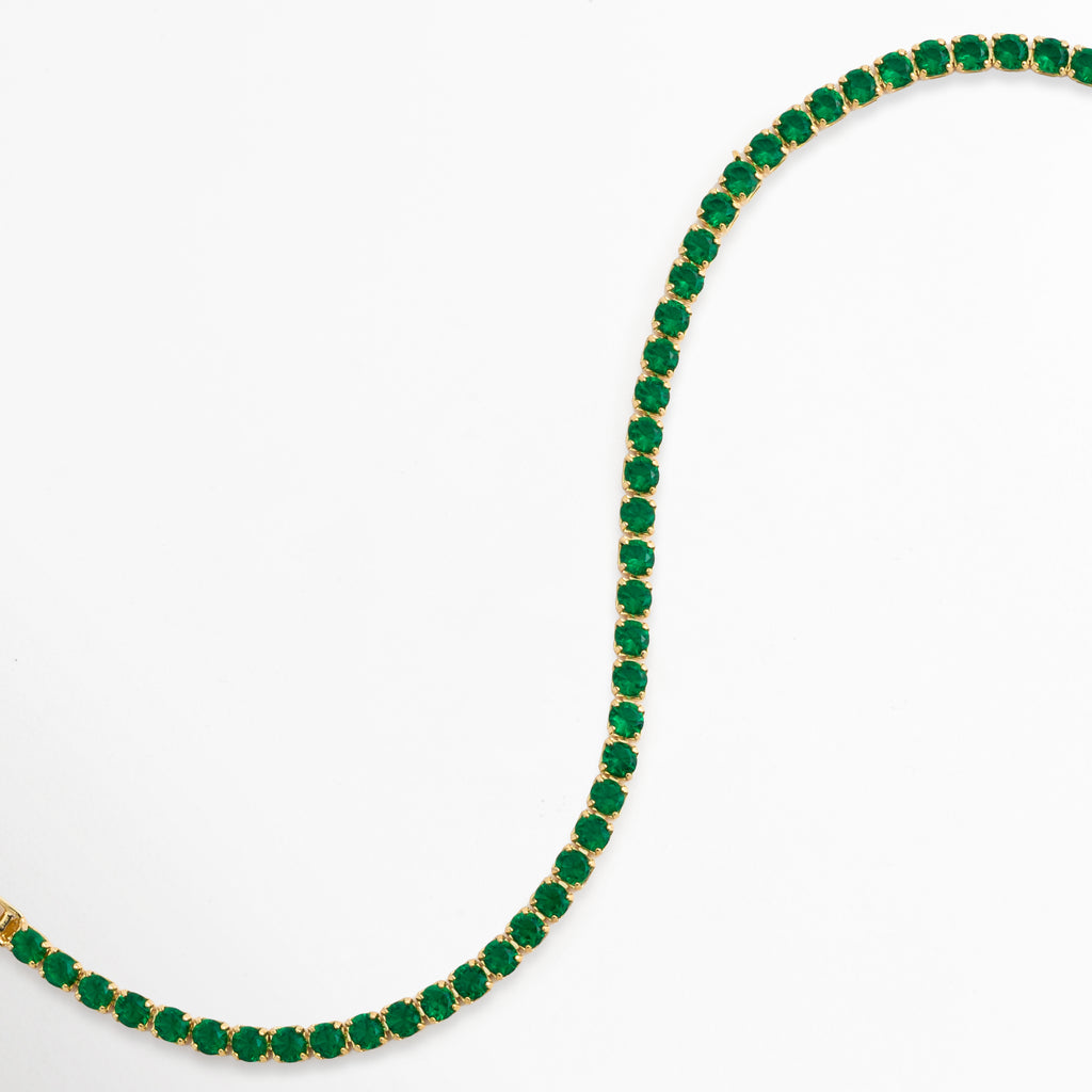 Vista en curva suave en S de la Tennis Bracelet Green Grace destacando la flexibilidad y el brillo de las circonias verdes, Acercamiento a los destellos de luz sobre la hilera de piedras tono esmeralda enchapadas en oro 18, Toma de perfil articulado que resalta la fluidez y caída de la pulsera tennis sobre fondo claro, Muestra de los reflejos cálidos del oro de 18K combinados con la intensidad de la pedrería verde. 