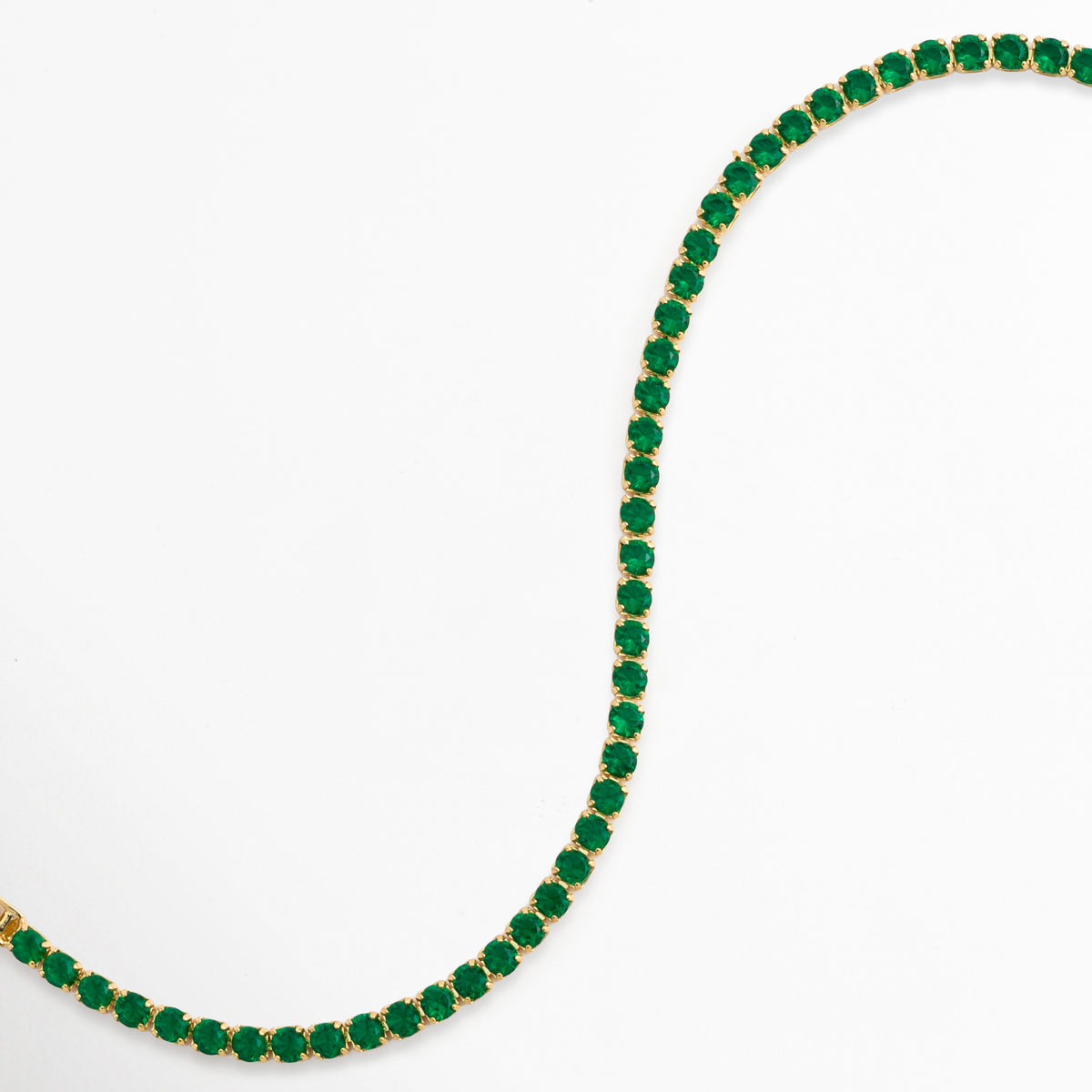 Vista en curva suave en S de la Tennis Bracelet Green Grace destacando la flexibilidad y el brillo de las circonias verdes, Acercamiento a los destellos de luz sobre la hilera de piedras tono esmeralda enchapadas en oro 18, Toma de perfil articulado que resalta la fluidez y caída de la pulsera tennis sobre fondo claro, Muestra de los reflejos cálidos del oro de 18K combinados con la intensidad de la pedrería verde. 