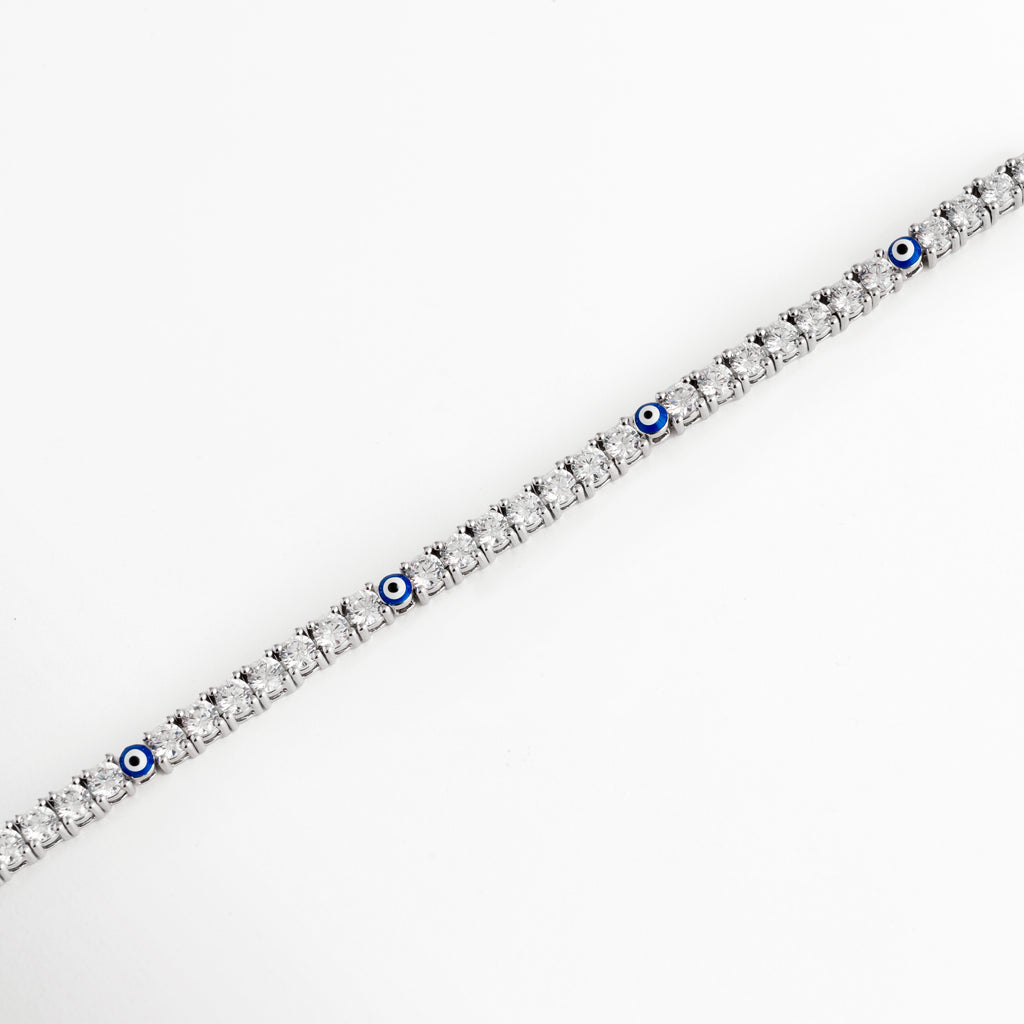 Vista en perspectiva de la pulsera tennis plateada mostrando la secuencia continua de circonias y los detalles intercalados de ojo turco azul, Acercamiento en ángulo a la estructura delgada de 0.3 cm con engaste de bisel/garras de pedrería fina enchapada en oro blanco, Toma en diagonal que resalta el brillo cristalino de las circonias transparentes y el contraste del esmalte azul del ojo turco, Muestra del flujo flexible y articulado de la pulsera tennis plateada