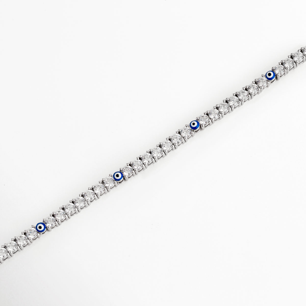 Vista en perspectiva de la pulsera tennis plateada mostrando la secuencia continua de circonias y los detalles intercalados de ojo turco azul, Acercamiento en ángulo a la estructura delgada de 0.3 cm con engaste de bisel/garras de pedrería fina enchapada en oro blanco, Toma en diagonal que resalta el brillo cristalino de las circonias transparentes y el contraste del esmalte azul del ojo turco, Muestra del flujo flexible y articulado de la pulsera tennis plateada