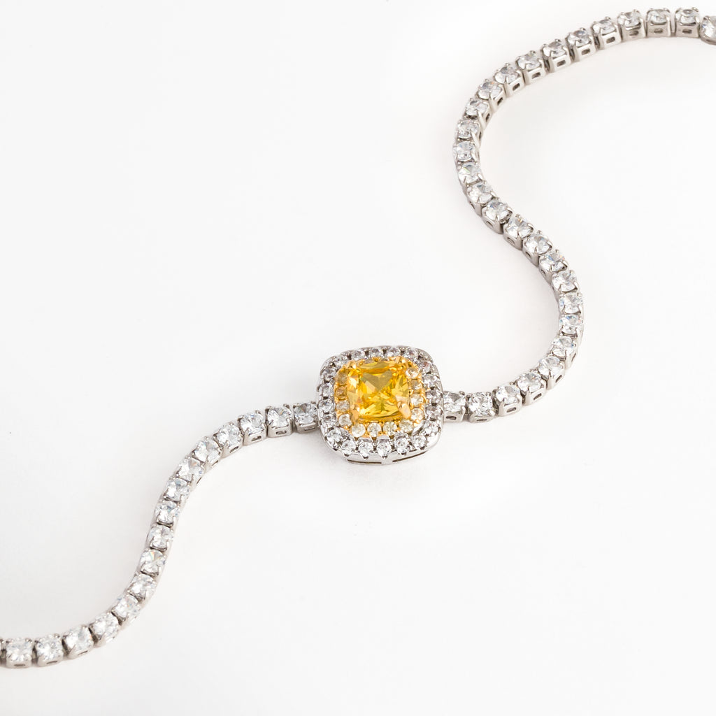 Vista en curva suave de la Pulsera Yellow destacando el brillo vibrante de la circonia central amarilla y el engaste del halo plateado, Acercamiento a la gema amarilla de corte cojín y la secuencia articulada de circonias transparentes de la pulsera tennis, Toma en diagonal que resalta el contraste entre la pedrería cristalina enchapada en oro blanco y el tono cálido de la piedra central, Muestra del flujo flexible de la cadena de circonias y la estructura