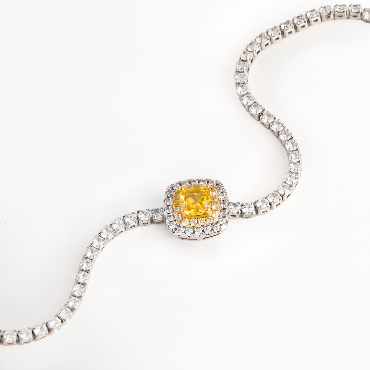 Vista en curva suave de la Pulsera Yellow destacando el brillo vibrante de la circonia central amarilla y el engaste del halo plateado, Acercamiento a la gema amarilla de corte cojín y la secuencia articulada de circonias transparentes de la pulsera tennis, Toma en diagonal que resalta el contraste entre la pedrería cristalina enchapada en oro blanco y el tono cálido de la piedra central, Muestra del flujo flexible de la cadena de circonias y la estructura