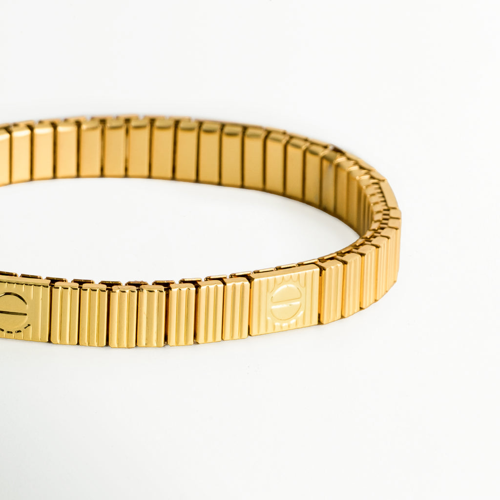 Pulsera Golden Vega vista en ángulo en primer plano, destacando los pliegues elásticos dorados y los grabados circulares en las placas, Vista de detalle de brazalete extensible dorado mostrando el acabado brillante de los eslabones acanalados y la bisagra flexible, Detalle de cerca de la textura acanalada y el grabado de tornillo o monograma en el brazalete elástico dorado, Toma inclinada de pulsera de acero enchapada en oro 18K con bandas verticales y placa decorativa.