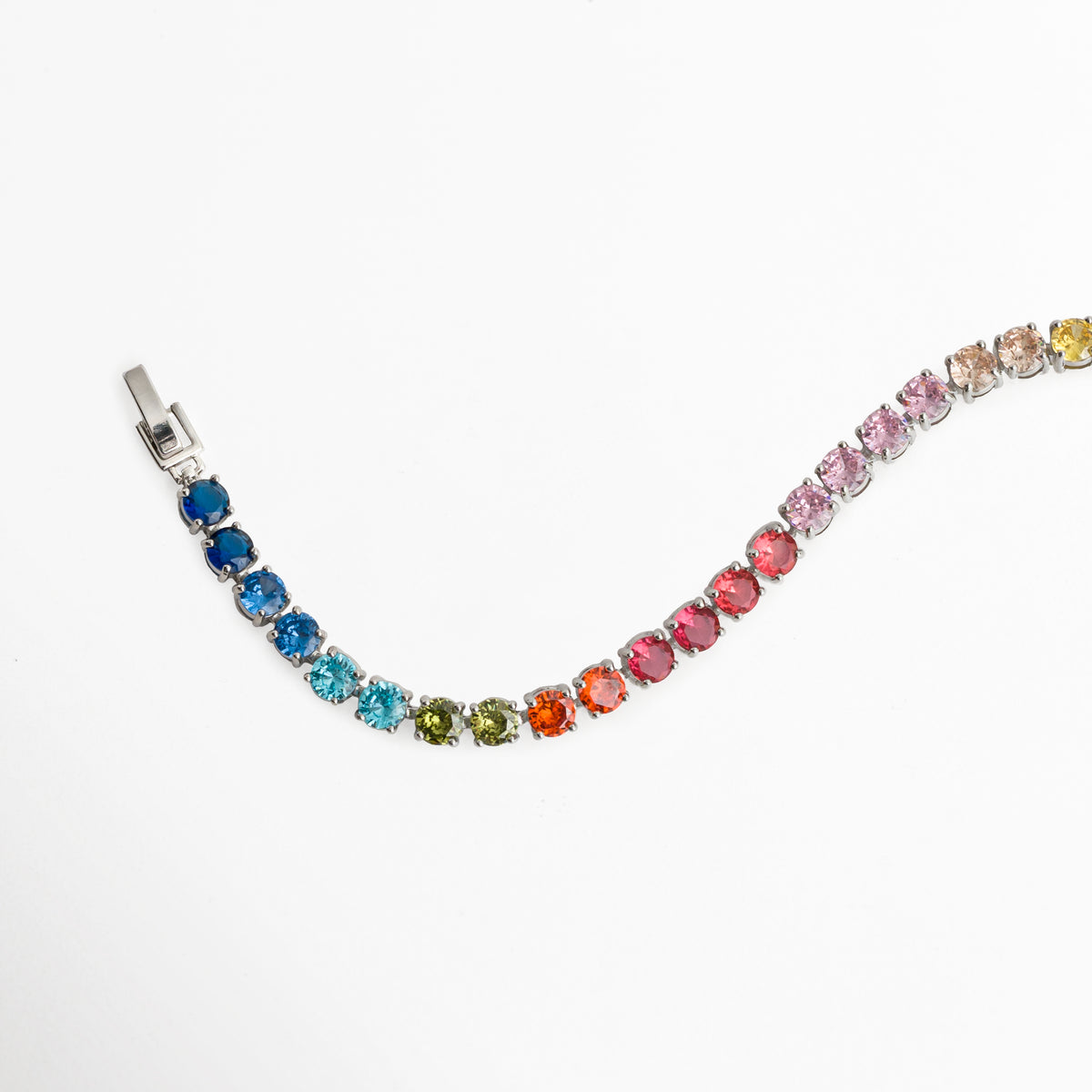 Pulsera Tennis Bracelet Rainbow dispuesta en una línea ondulada suave mostrando la flexibilidad del engaste de circonias, Vista en perspectiva de la pulsera tennis multicolor desplegada en curva sobre fondo blanco, Muestra del degradé cromático continuo desde el rojo vibrante hasta el azul profundo en la pulsera enchapada en rodio, Detalle de la articulación flexible y el brillo cristalino de las gemas de colores de 0.5 cm de ancho.