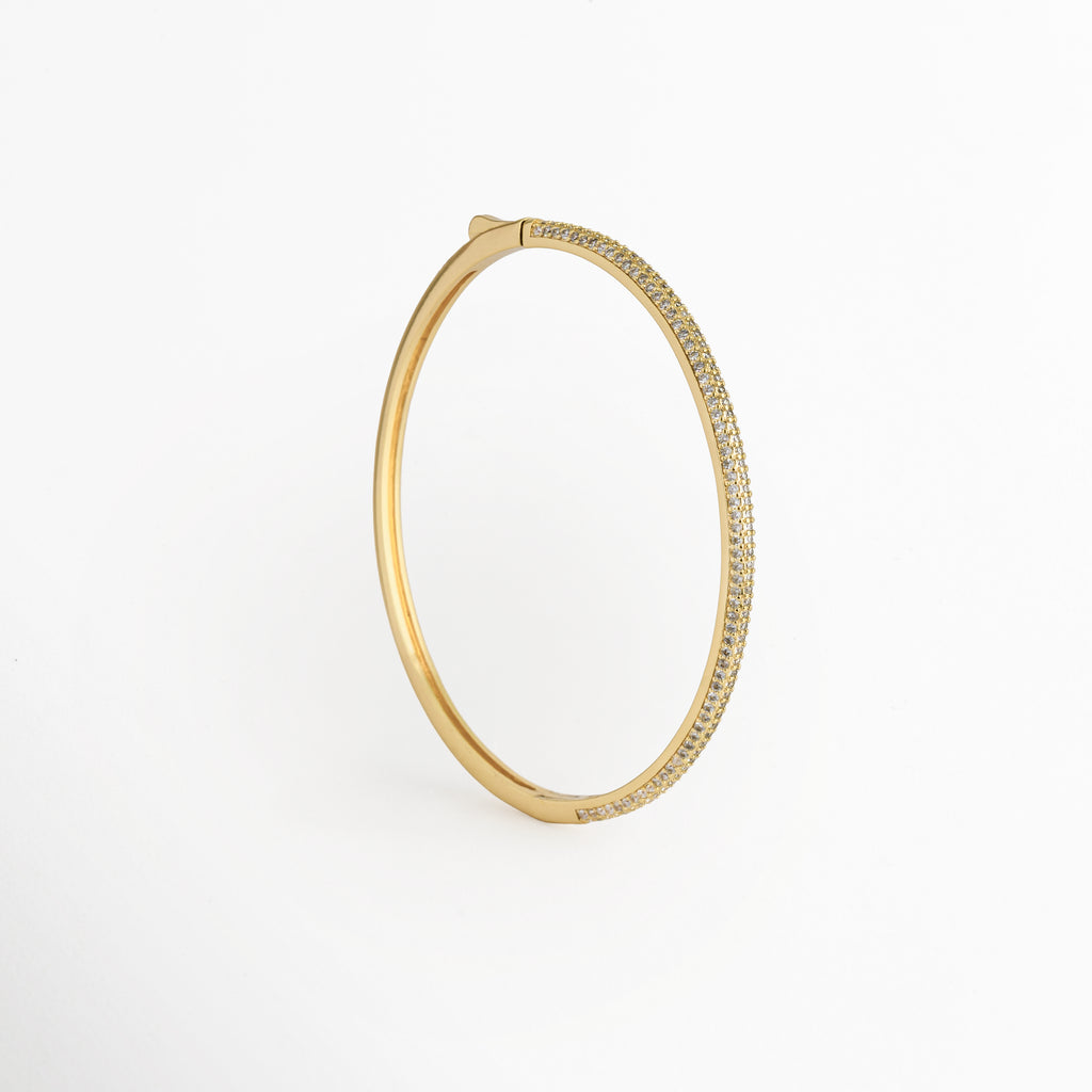 Vista aérea en posición vertical de la Pulsera Golden Liore resaltando su forma ovalada ergonómica, Toma cenital del brazalete rígido dorado mostrando el contraste entre la cara pavé de circonias y el broche superior, Plano general vertical de pulsera tipo bangle de 0.3 cm de grosor sobre fondo blanco neutro, Muestra del contorno limpio y redondeado de la Pulsera Golden Liore en tono oro cálido.  