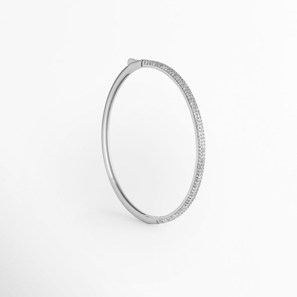 Vista aérea en posición vertical de la Pulsera Silver Liore resaltando su forma ovalada ergonómica, Toma cenital del brazalete rígido plateado mostrando el contraste entre la cara pavé de circonias y el broche superior, Plano general vertical de pulsera tipo bangle de 0.3 cm de grosor sobre fondo blanco neutro, Muestra del contorno limpio y redondeado de la Pulsera Silver Liore en tono oro blanco frío.  