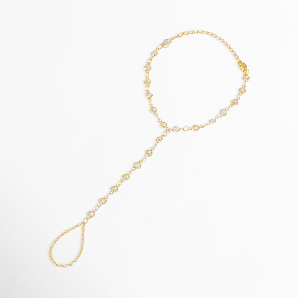 Pulsera Esclava Zayra de cobre enchapada en oro de 18 cm con extensión de 5 cm, con hileras de circonias biceladas que conectan la muñeca al dedo, vista frontal sobre fondo blanco, Vista de frente de pulsera de mano o esclava dorada tipo hand chain con circonias redondas transparentes engastadas en bisel, Plano general de esclava de mano dorada con anillo de cadena integrado e incrustaciones de pedrería cristalina sobre fondo neutro.