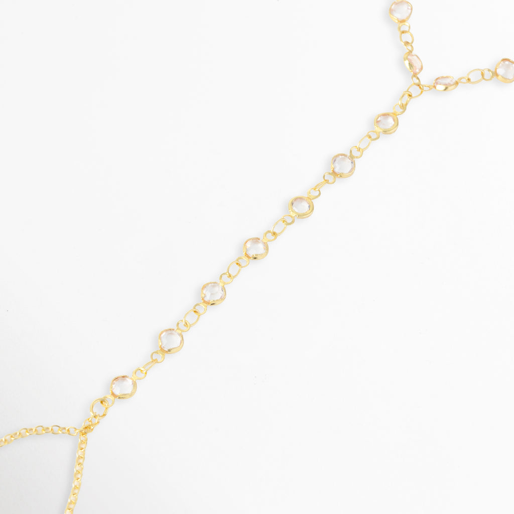 Acercamiento a la secuencia de circonias engastadas en bisel que recorren el dorso de la mano hasta el anillo de cadena en la Pulsera Esclava Zayra, Vista en perspectiva de la unión entre la tira lineal con pedrería y la lazada dorada para el dedo, Toma de detalle que resalta la claridad de los cristales redondos de corte brillante y el acabado enchapado en oro, Muestra del trabajo de engaste individual en cada eslabón con circonia sobre fondo blanco.
