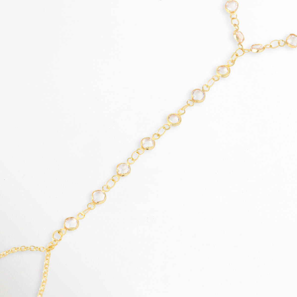 Acercamiento a la secuencia de circonias engastadas en bisel que recorren el dorso de la mano hasta el anillo de cadena en la Pulsera Esclava Zayra, Vista en perspectiva de la unión entre la tira lineal con pedrería y la lazada dorada para el dedo, Toma de detalle que resalta la claridad de los cristales redondos de corte brillante y el acabado enchapado en oro, Muestra del trabajo de engaste individual en cada eslabón con circonia sobre fondo blanco.