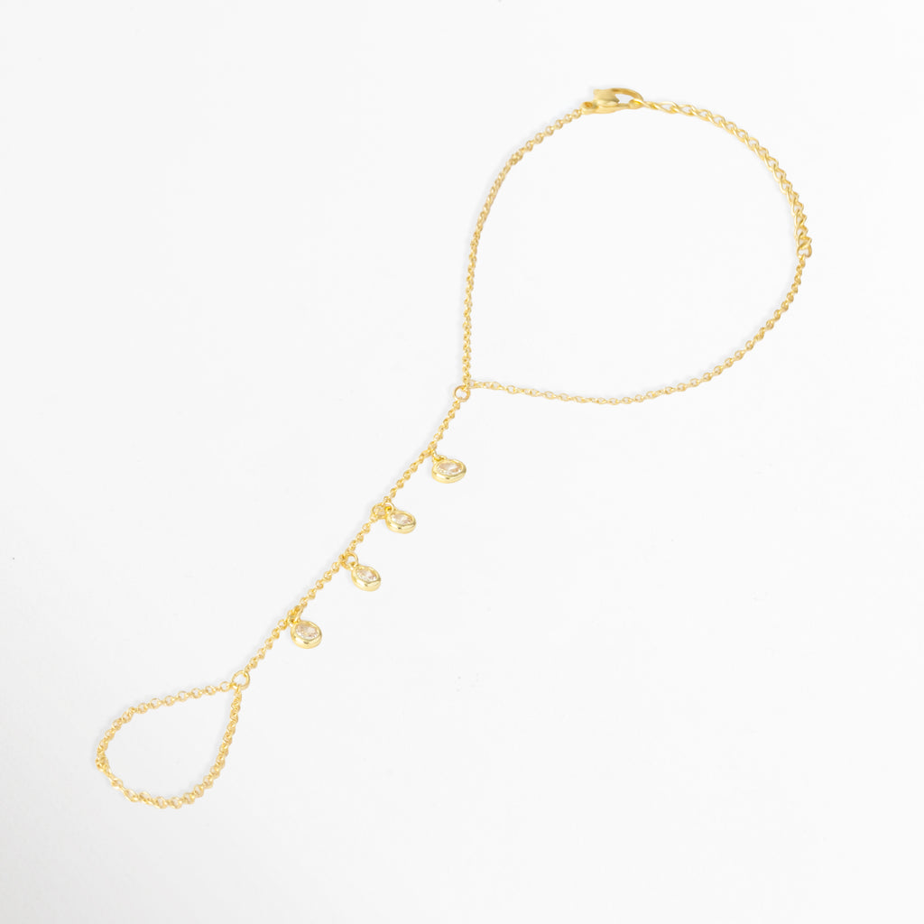 Pulsera Esclava Chita de cobre enchapada en oro de 17 cm con extensión de 5 cm, con charms colgantes de circonia en la tira dorsal conectada al dedo, vista frontal sobre fondo blanco, Vista de frente de pulsera de mano o esclava dorada tipo hand chain con pequeños dijes de circonia transparente que cuelgan del dorso, Plano general de esclava de mano dorada con hileras de charms de circonia brillante y anillo de cadena integrado sobre fondo neutro.
