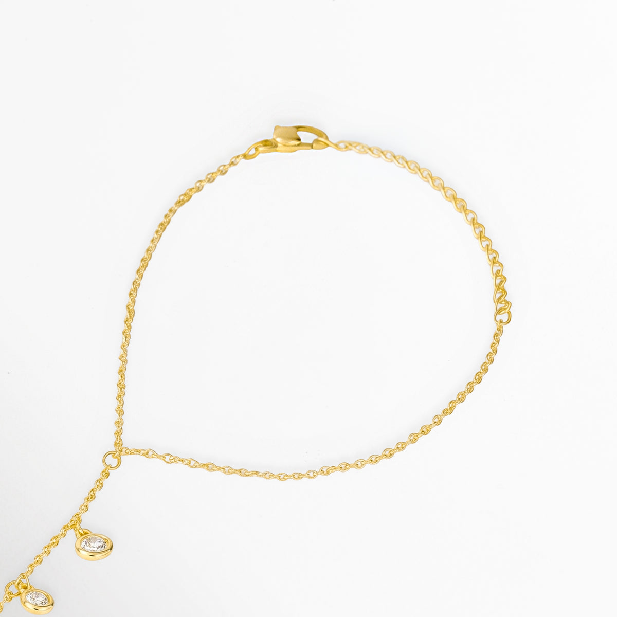 Acercamiento al punto de unión entre la cadena de muñeca y la tira dorsal con charms colgantes en la Pulsera Esclava Chita, Vista en ángulo del broche de mosquetón y la caída de los primeros dijes de circonia sobre la cadena dorada, Toma de detalle que muestra la fluidez de la cadena base de cobre enchapada en oro de alto brillo, Muestra del engaste metálico y el brillo de las circonias transparentes suspendidas sobre fondo blanco.