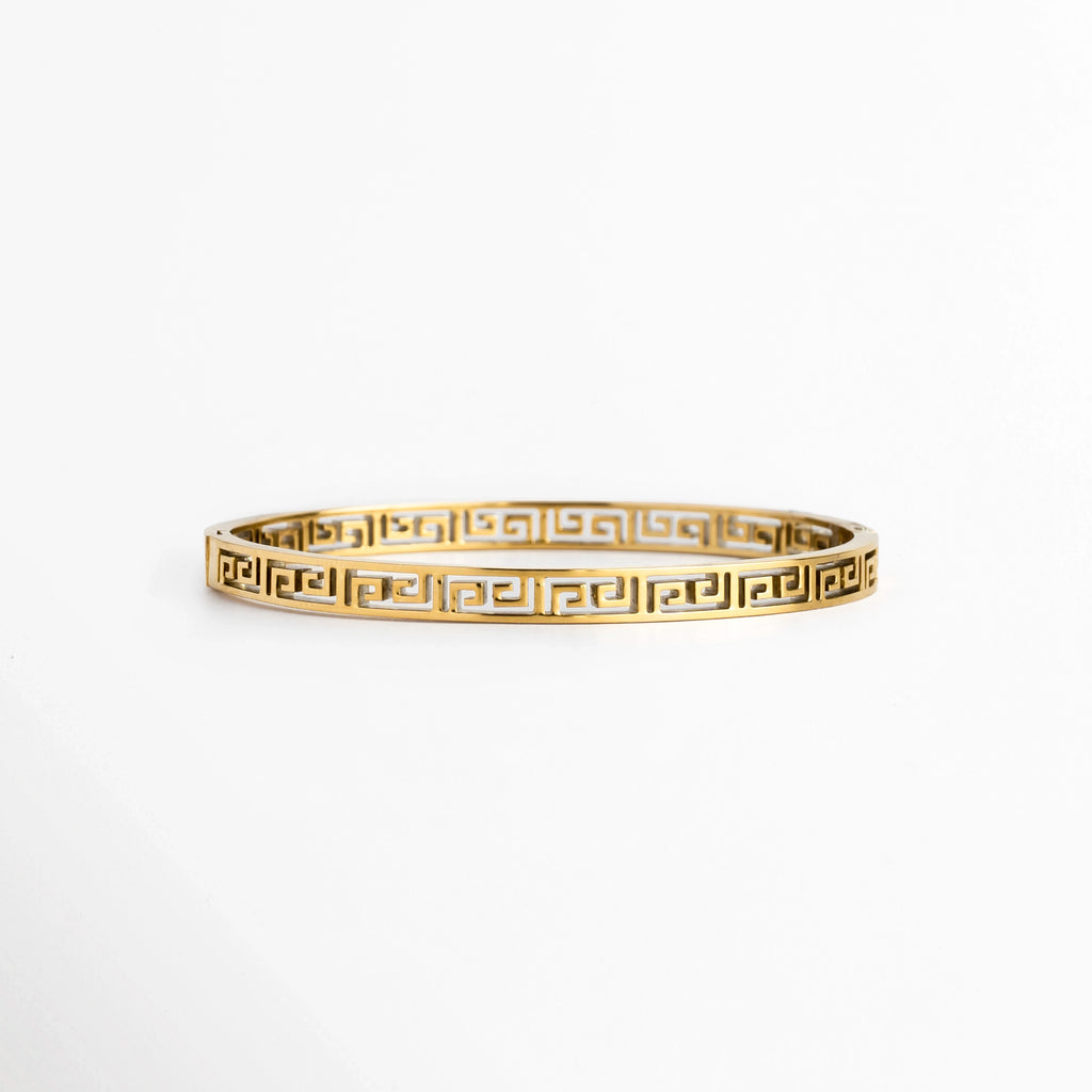 Pulsera Amani de acero de titanio enchapada en oro con diseño rígido de greca clásica o meandro calado de 0.5 cm de ancho, vista frontal sobre fondo blanco, Vista de frente de brazalete dorado de silueta ovalada con patrón geométrico estilo greca griega en calado continuo, Plano general de pulsera rígida en tono oro brillante con estructura arquitectónica de líneas labradas sobre fondo neutro, Pulsera Amani destacando el acabado de alto brillo sobre el acero