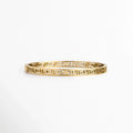Pulsera Amani de acero de titanio enchapada en oro con diseño rígido de greca clásica o meandro calado de 0.5 cm de ancho, vista frontal sobre fondo blanco, Vista de frente de brazalete dorado de silueta ovalada con patrón geométrico estilo greca griega en calado continuo, Plano general de pulsera rígida en tono oro brillante con estructura arquitectónica de líneas labradas sobre fondo neutro, Pulsera Amani destacando el acabado de alto brillo sobre el acero