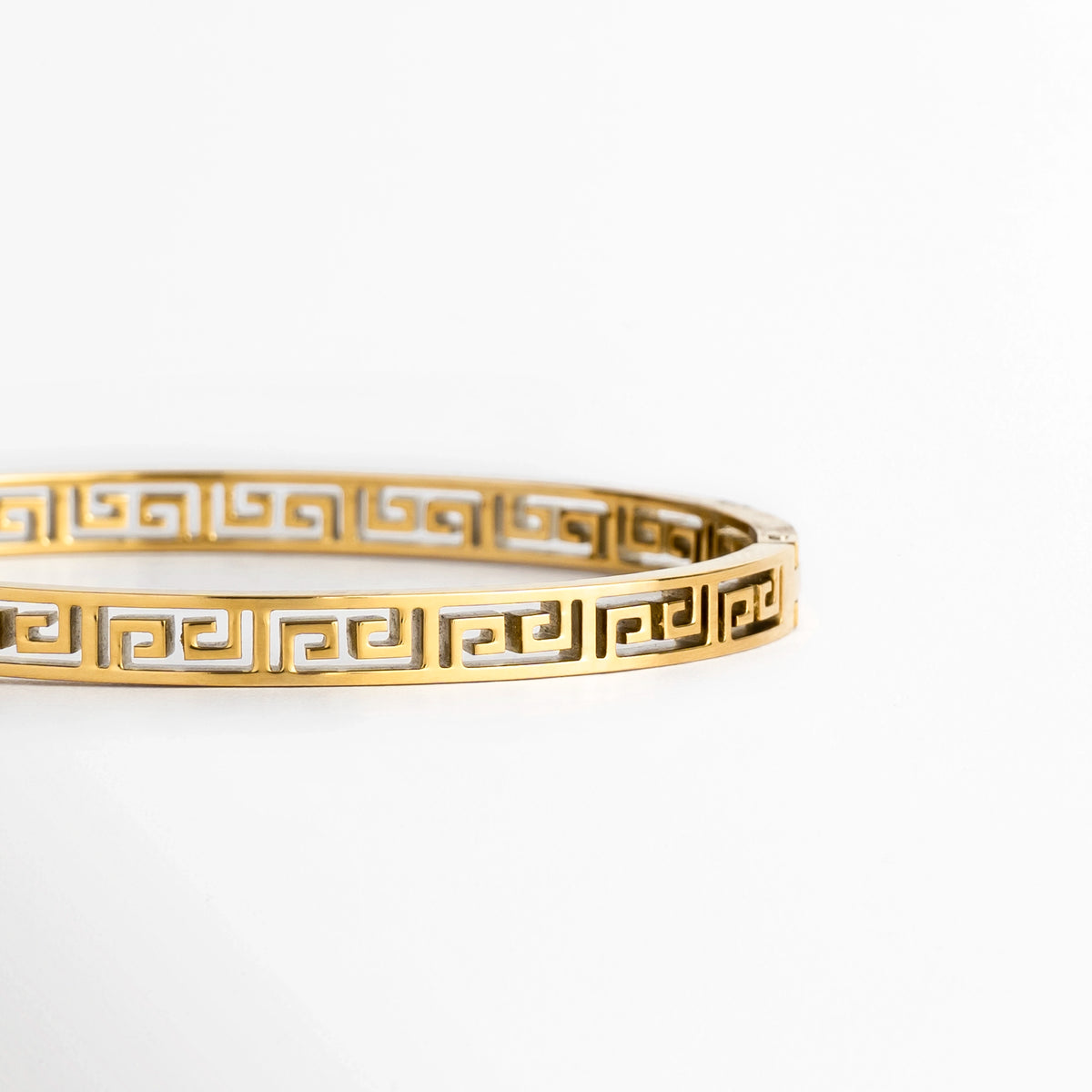 Acercamiento lateral al calado geométrico en meandro o greca clásica de la Pulsera Amani enchapada en oro, Toma de perfil en ángulo que resalta la nitidez de los bordes y los espacios calados en la caña metálica de 0.5 cm, Muestra del pulido espejo y la solidez del acero de titanio dorado sobre superficie blanca, Plano detalle de la repetición del motivo grecolatino en la cara exterior del brazalete rígido.