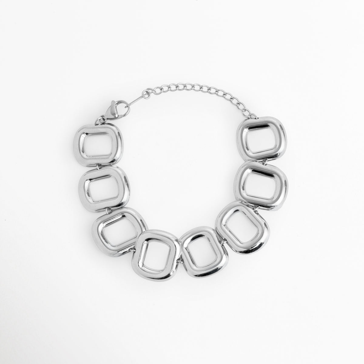 Pulsera Freire de acero inoxidable enchapada en oro blanco con eslabones geométricos cuadrados de bordes redondeados y silueta abombada, vista superior sobre fondo blanco, Vista frontal de brazalete plateado con eslabones calados cuadrados de 2.3 cm de ancho y cadena de extensión ajustable, Plano general de pulsera gruesa de eslabones estructurales abombados en acabado oro blanco sobre fondo neutro, Pulsera Freire destacando la simetría de sus eslabones huecos