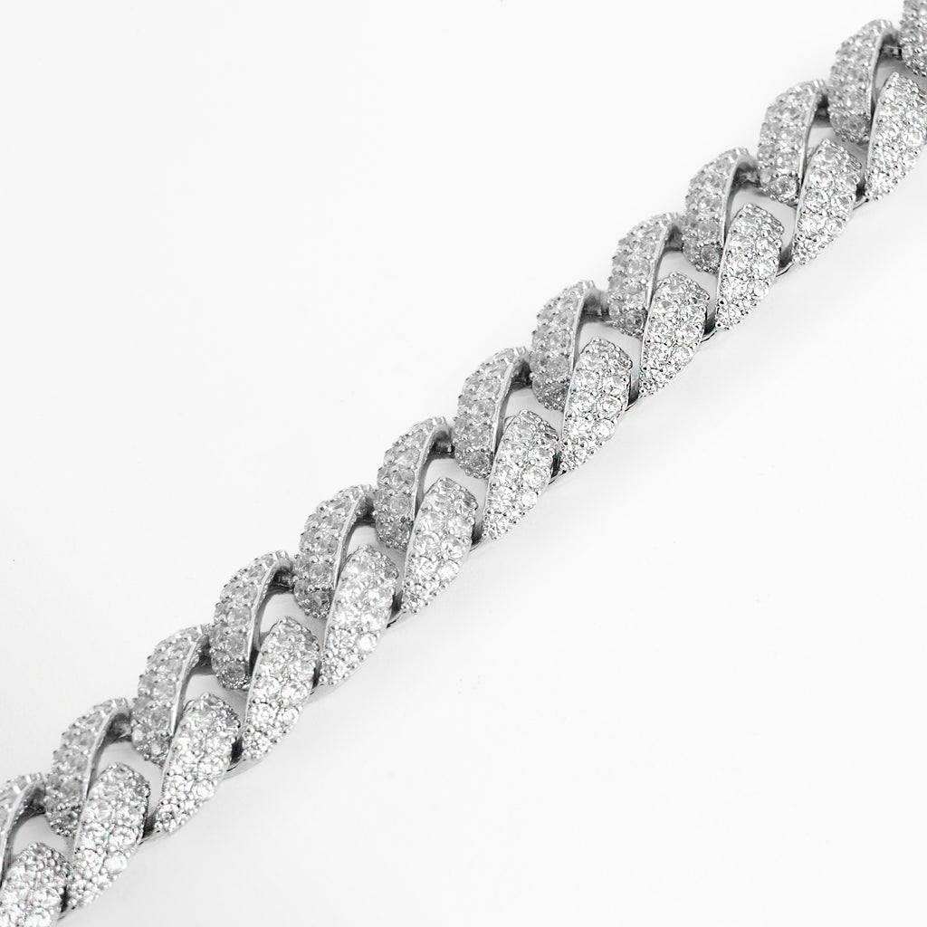 Detalle en ángulo de los eslabones entrelazados de la pulsera Oriana evidenciando la densidad del pavé de circonias en tono oro blanco, Acercamiento diagonal a la textura iced out de los eslabones barbados planos de 1 cm de ancho, Vista de cerca del brillo cristalino y engaste de precisión de las circonias sobre el tejido de la cadena cubana, Toma en perspectiva de la superficie cuajada de piedras transparentes sobre el acabado enchapado en oro blanco.