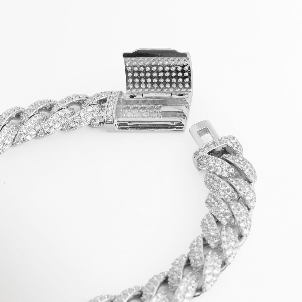 Pulsera Oriana con el broche de cajón con doble seguro lateral desplegado, mostrando el mecanismo de cierre iced out, Vista de cerca del broche rectangular cuajado de circonias abierto con su lengüeta de encaje para pulsera cubano, Detalle del sistema de sujeción de alta seguridad enchapado en oro blanco en toma diagonal sobre fondo blanco, Muestra del mecanismo de cierre de cajón con pavé de pedrería de la pulsera de eslabones de 1 cm.