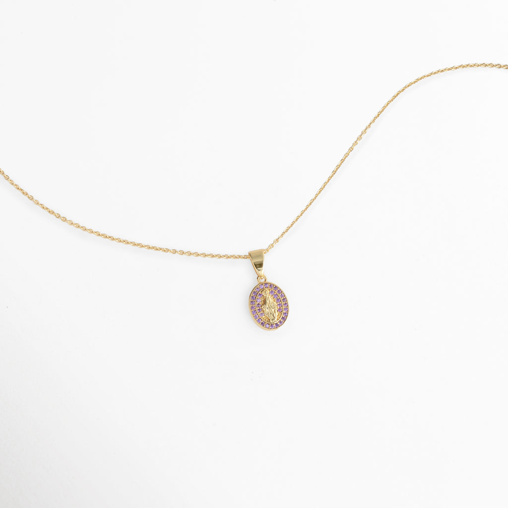 Vista aérea en curva suave del Collar Medallón Dorado Virgen con el dije ovalado de 1.5 cm de ancho reposando sobre fondo blanco, Toma extendida de la fina cadena dorada de 47 cm mostrando la proporción de la medalla religiosa con circonias violeta, Muestra de la silueta delicada de la cadena y el resplandor de la pedrería púrpura en ángulo horizontal,  Acercamiento a la caída ligera del collar religioso de tono dorado sobre superficie clara