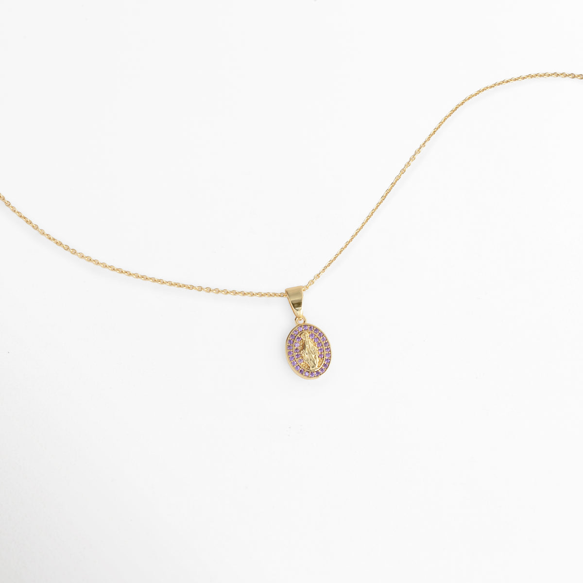 Vista aérea en curva suave del Collar Medallón Dorado Virgen con el dije ovalado de 1.5 cm de ancho reposando sobre fondo blanco, Toma extendida de la fina cadena dorada de 47 cm mostrando la proporción de la medalla religiosa con circonias violeta, Muestra de la silueta delicada de la cadena y el resplandor de la pedrería púrpura en ángulo horizontal,  Acercamiento a la caída ligera del collar religioso de tono dorado sobre superficie clara