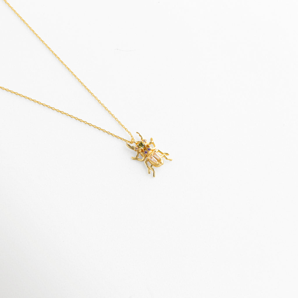 Vista inclinada de Collar Beetle con dije de escarabajo y zirconias brillantes, Detalle del colgante de escarabajo dorado con acabado sofisticado y femenino, Collar con dije Beetle en vista diagonal y brillo elegante, Cadena fina dorada con colgante de escarabajo decorado con zirconias, Detalle de collar delicado con dije místico de escarabajo en tono dorado.