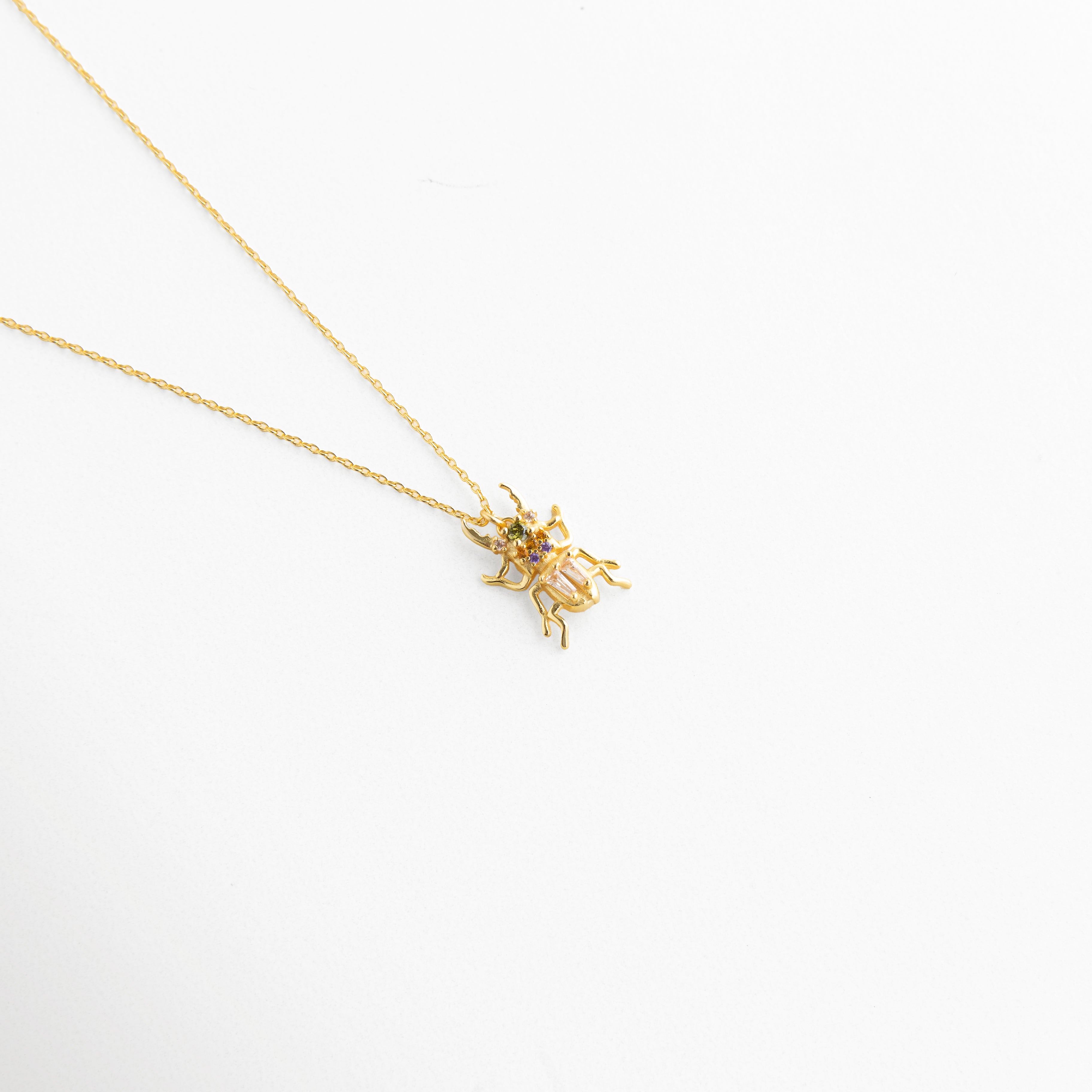 Vista inclinada de Collar Beetle con dije de escarabajo y zirconias brillantes, Detalle del colgante de escarabajo dorado con acabado sofisticado y femenino, Collar con dije Beetle en vista diagonal y brillo elegante, Cadena fina dorada con colgante de escarabajo decorado con zirconias, Detalle de collar delicado con dije místico de escarabajo en tono dorado.