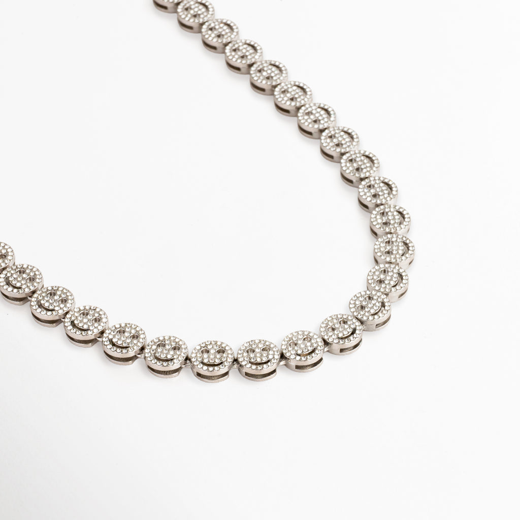 Detalle en ángulo del collar Felissia mostrando la alineación de las caritas felices, Vista tridimensional de la cadena plateada con zirconias brillantes, Enfoque lateral del grosor y acabado pulido en oro blanco del collar, Sucesión de eslabones redondos con diseño sonriente y pedrería fina, Primer plano de la textura y el patrón reflectante del collar con diseño aesthetic.