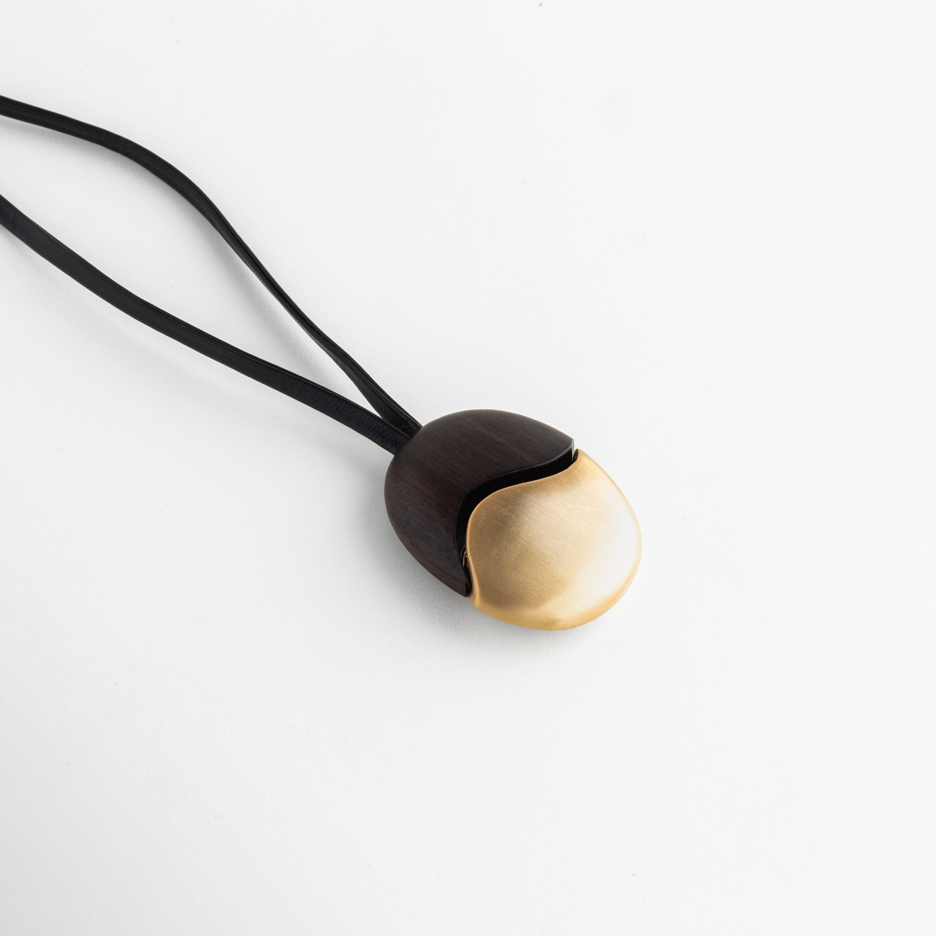 Collar Gold Monet con dije de madera de ébano y baño de oro 18K sobre fondo blanco, Detalle de collar artesanal: contraste de madera oscura y metal dorado satinado, Joyería de autor: collar de cuero negro con dije bimetálico ovalado en oro y ébano, Vista de cerca de collar minimalista con terminales doradas y cordón de cuero doble, Accesorio de Collar Gold Monet con dije de madera de ébano y baño de oro 18K sobre fondo blanco