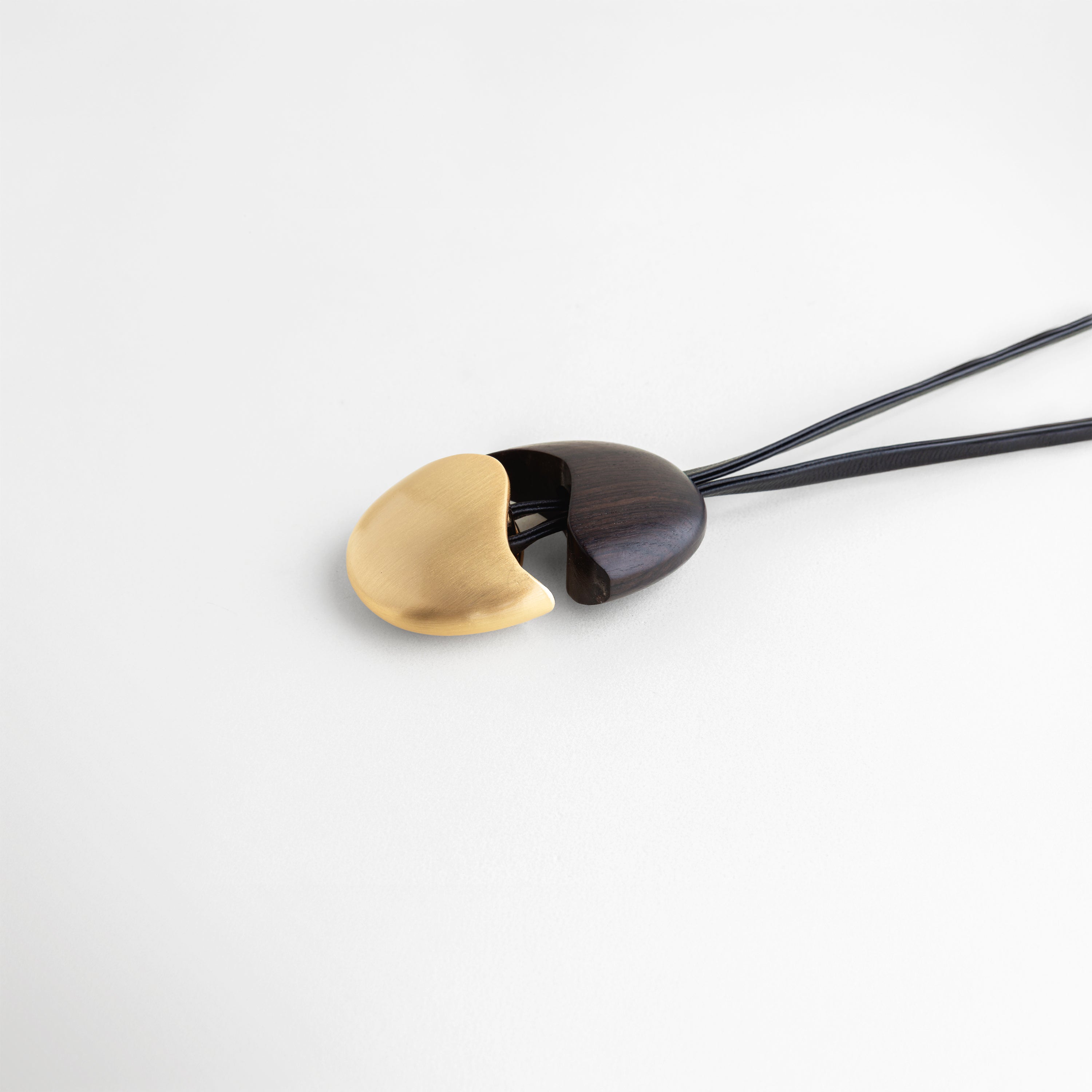 Vista angular del dije Gold Monet resaltando el brillo del oro sobre la madera, Dije orgánico en posición inclinada con cordón de cuero negro y herrajes dorados, Perspectiva cenital de joyería contemporánea en madera de ébano y oro de 18K, Unión curva perfecta entre madera negra y metal dorado en dije de diseño oval, Composición minimalista de collar artístico con acabados pulidos y cordón ajustable.