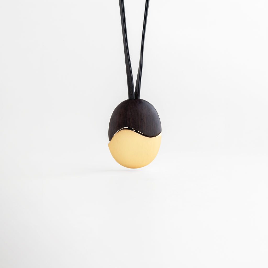Dije Gold Monet recostado mostrando el relieve y la transición ébano-oro, Vista de perfil bajo del dije ovalado con acabado enchapado en oro y madera, Detalle lateral de la pieza central resaltando su forma ergonómica y pulido, Fotografía de producto: collar con dije bimetálico dorado en reposo sobre blanco, Diseño escultural: transición orgánica entre madera negra y oro en el dije Monet.