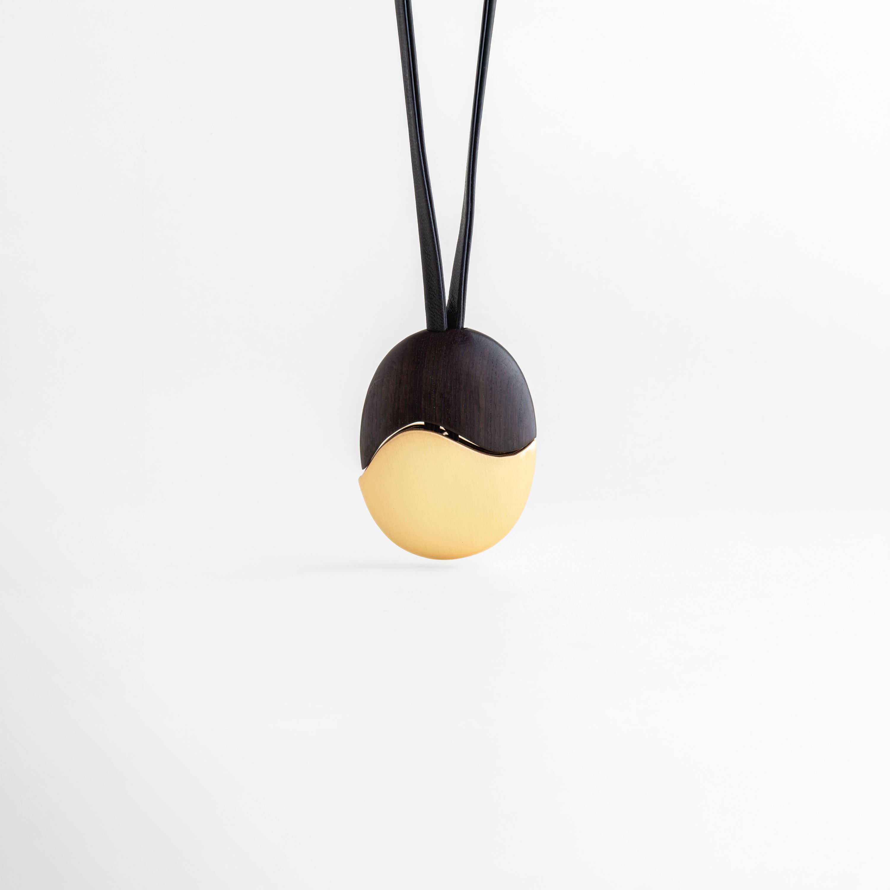 Dije Gold Monet recostado mostrando el relieve y la transición ébano-oro, Vista de perfil bajo del dije ovalado con acabado enchapado en oro y madera, Detalle lateral de la pieza central resaltando su forma ergonómica y pulido, Fotografía de producto: collar con dije bimetálico dorado en reposo sobre blanco, Diseño escultural: transición orgánica entre madera negra y oro en el dije Monet.