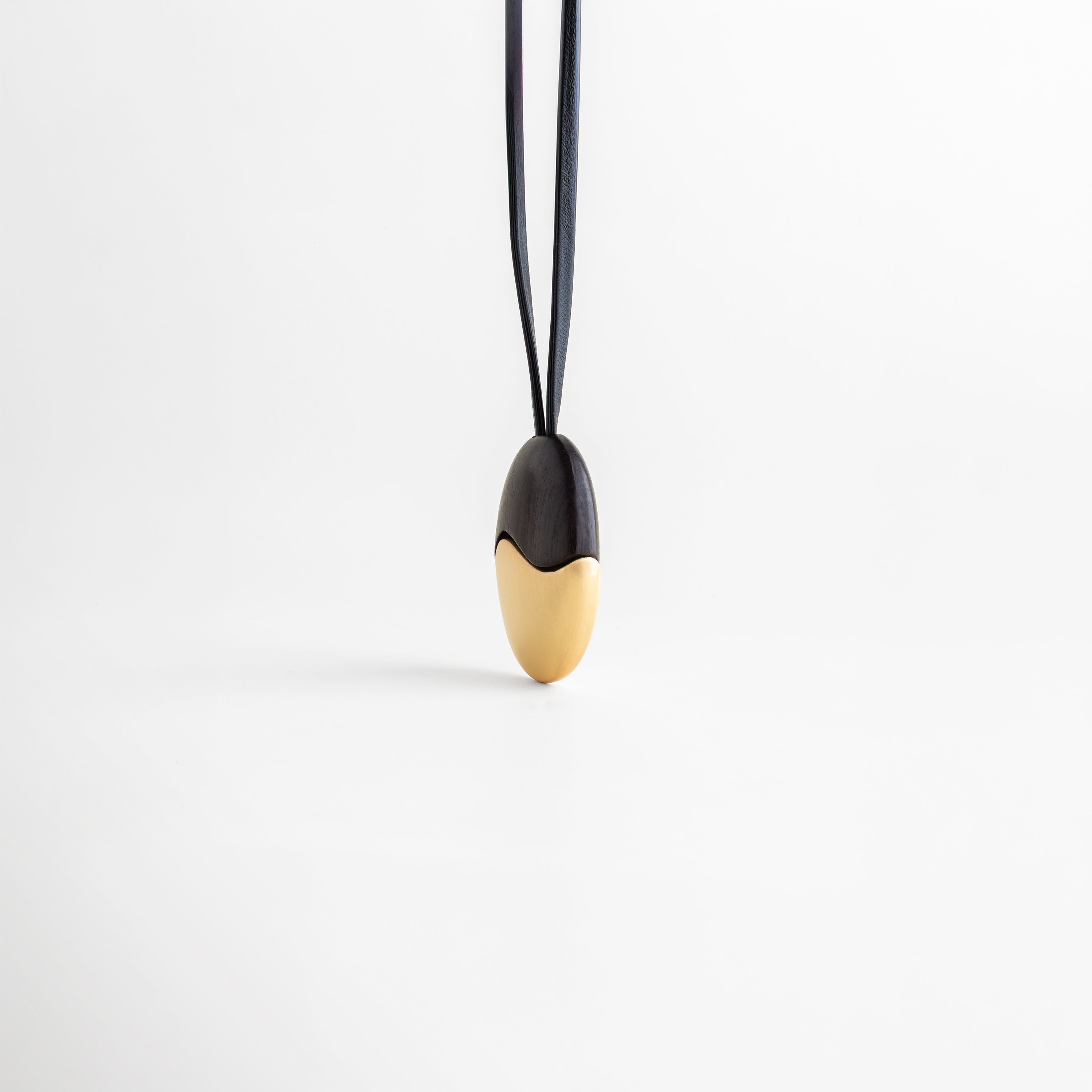 Collar Gold Monet colgando verticalmente con equilibrio entre ébano y oro, Vista frontal de dije ovalado dividido simétricamente en madera y metal dorado, Collar minimalista suspendido que muestra la caída elegante del cordón de cuero, Diseño de inspiración artística: collar de ébano y oro estilo Monet vista frontal, Contraste de alta gama: collar de cuero con dije dorado y madera para mujer.