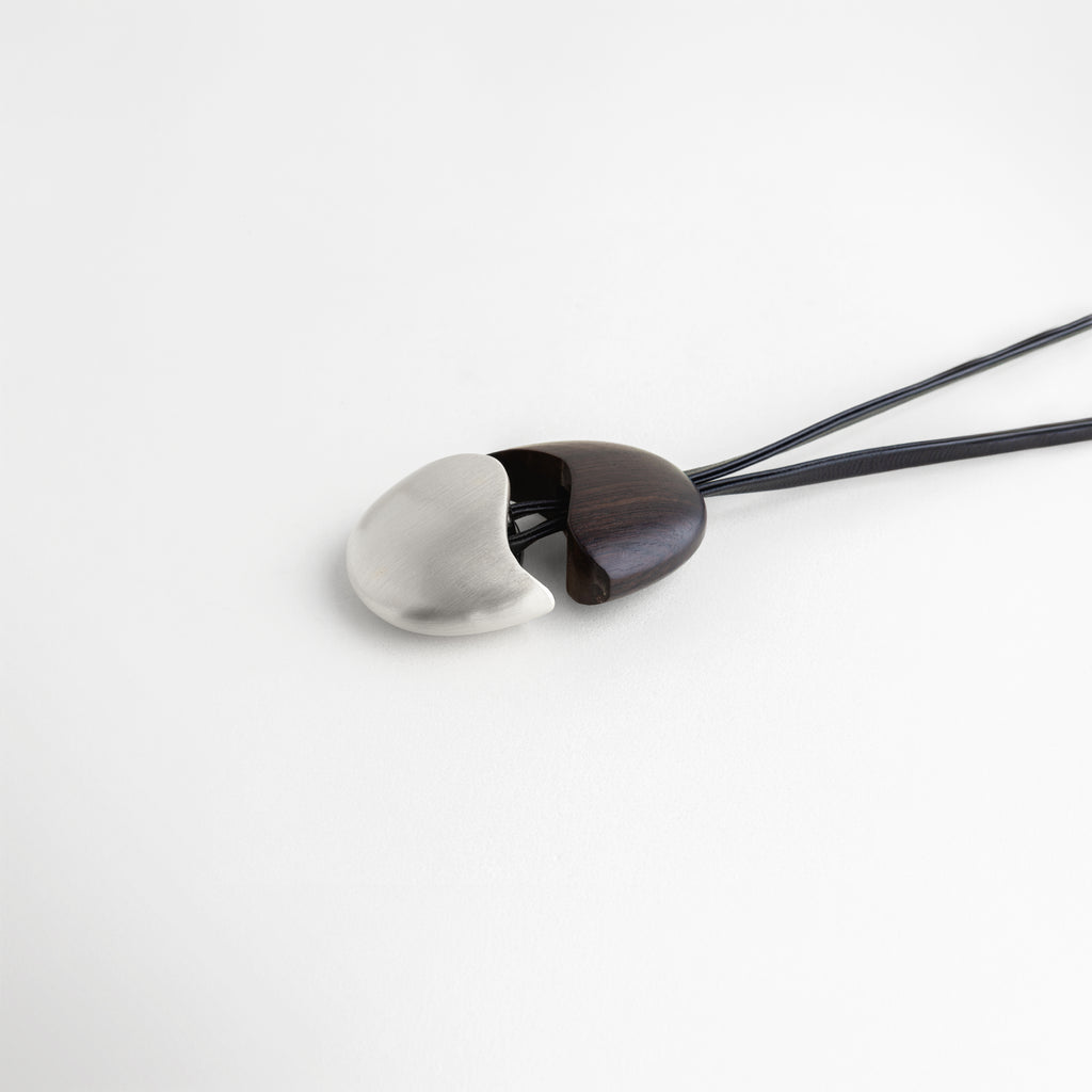 Dije Silver Monet recostado mostrando la unión curva entre el ébano y el acero, Vista de perfil bajo del dije ovalado con acabado enchapado en acero y madera, Detalle lateral de la pieza central del collar resaltando su forma ergonómica, Fotografía de producto: collar con dije bimetálico en reposo sobre superficie blanca, Diseño escultural: transición suave entre madera negra y acero en el dije Monet.