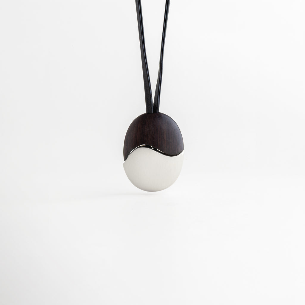 Collar Silver Monet colgando verticalmente con equilibrio entre madera y metal, Vista frontal de dije ovalado dividido simétricamente en ébano y acero plateado, Collar minimalista suspendido que muestra la caída del cordón de cuero negro, Diseño de inspiración artística: collar de ébano y acero estilo Monet frontal, Simetría orgánica en collar de cuero con dije de alto contraste para mujer.