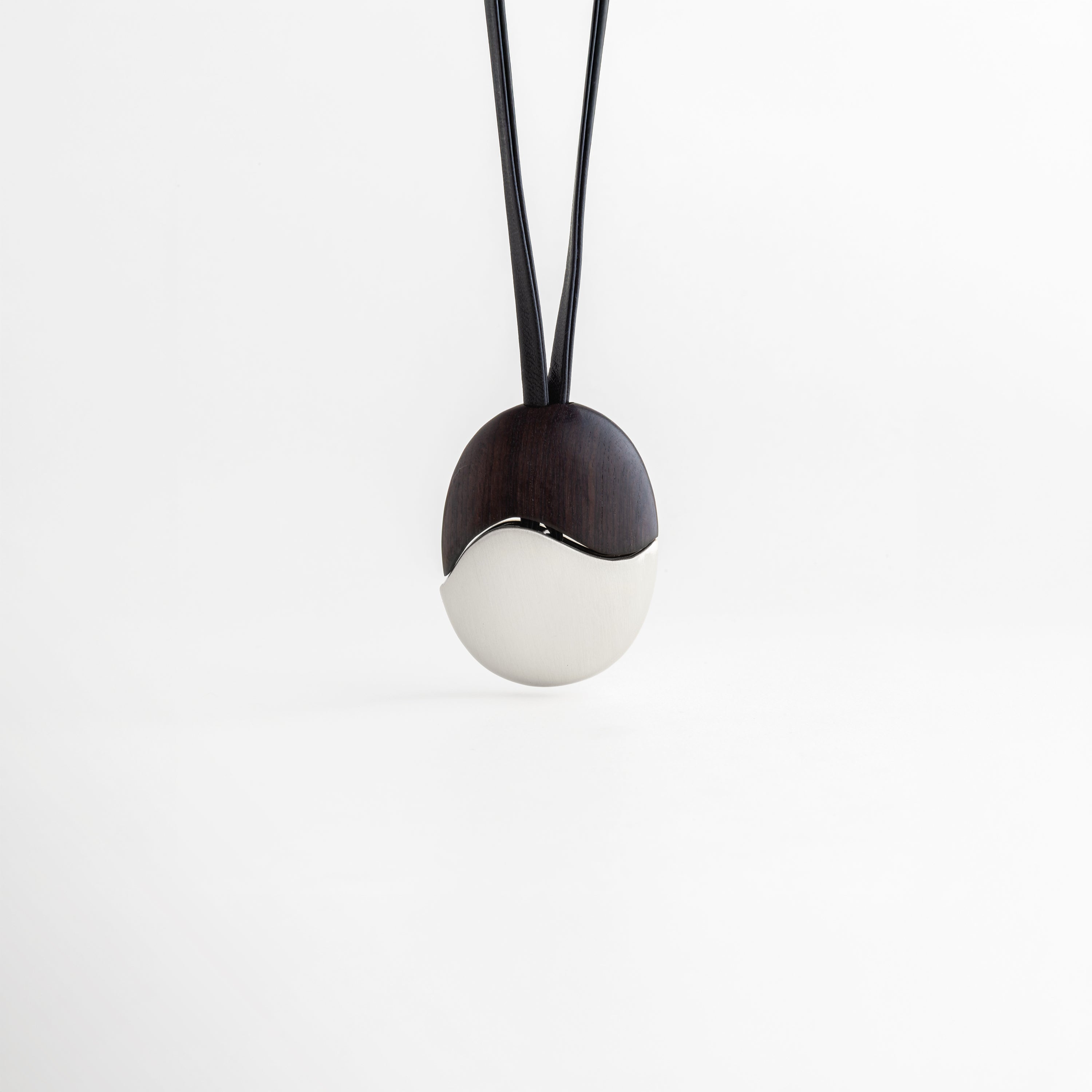 Collar Silver Monet colgando verticalmente con equilibrio entre madera y metal, Vista frontal de dije ovalado dividido simétricamente en ébano y acero plateado, Collar minimalista suspendido que muestra la caída del cordón de cuero negro, Diseño de inspiración artística: collar de ébano y acero estilo Monet frontal, Simetría orgánica en collar de cuero con dije de alto contraste para mujer.