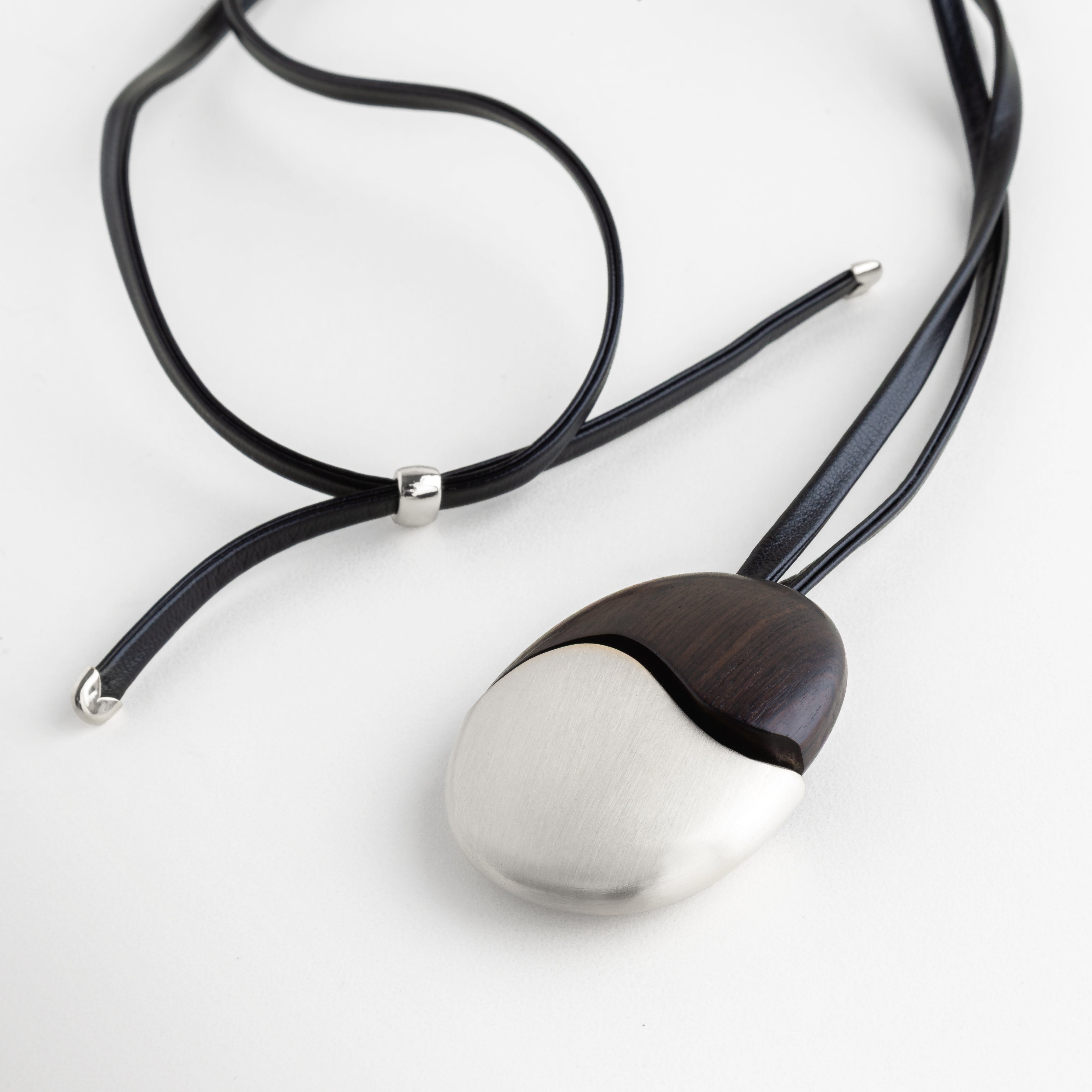 Collar Silver Monet con dije ovalado de madera de ébano y acero sobre fondo blanco, Detalle de collar artesanal con contraste de madera oscura y metal plateado mate, Joyería contemporánea: collar de cuero negro con dije bimetálico de ébano y acero, Vista cercana de collar minimalista con terminales metálicas y cordón de cuero, Accesorio de diseño: collar Monet con acabado en acero enchapado y madera noble.