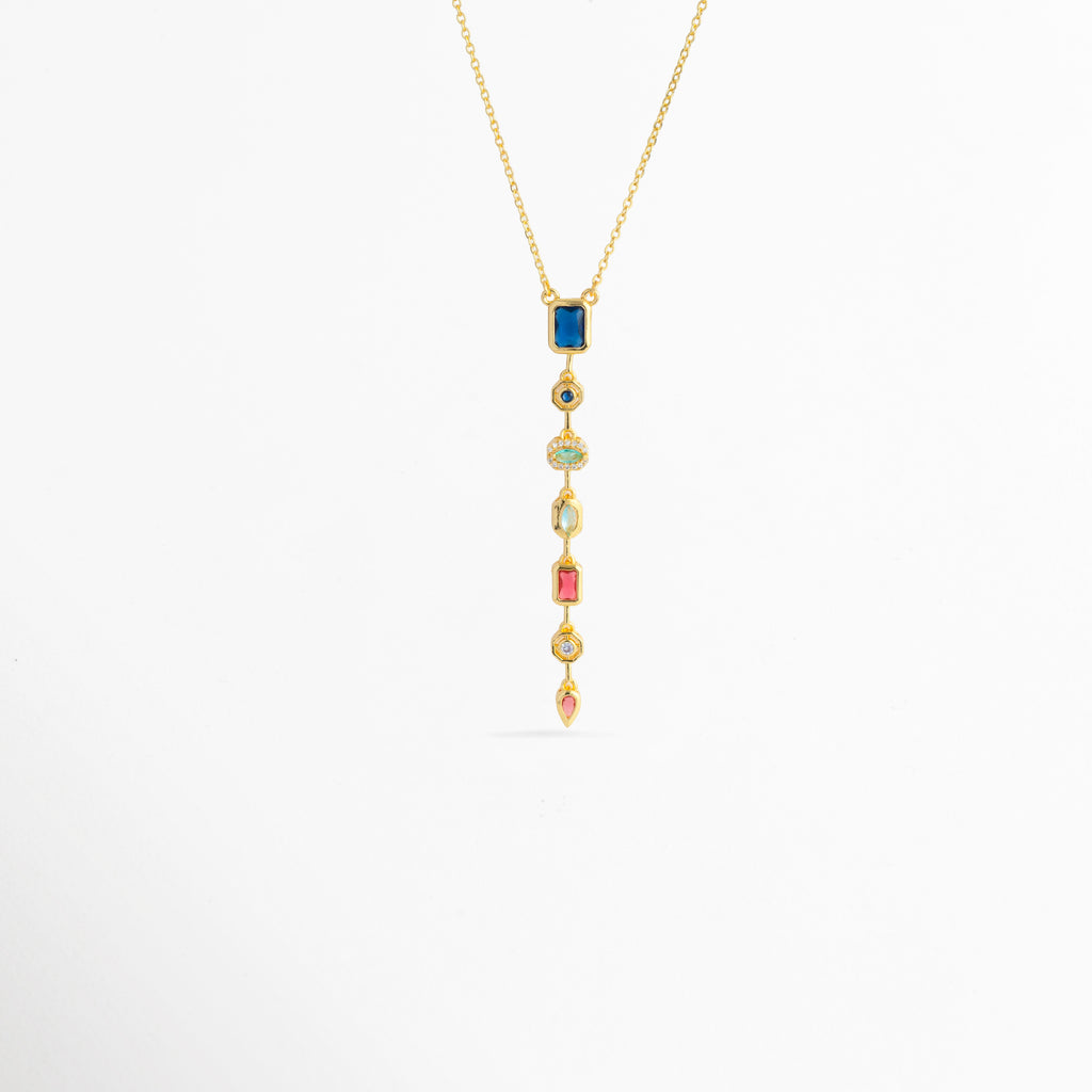 Collar Prisma Dorado enchapado en oro 18K con dije colgante lineal vertical compuesto por siete circonias de colores en diferentes cortes, Vista de frente de collar dorado con colgante alargado estilo cascada de gemas multicolor sobre fondo blanco, Dije vertical de 7.5 cm con gemas de tonos azul, turquesa, fucsia, amarillo y rosa suspendido de una fina cadena dorada, Collar Prisma Dorado con composición geométrica de circonias multicolores enmarcadas en biseles