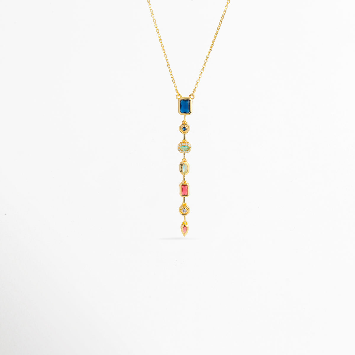 Collar Prisma Dorado enchapado en oro 18K con dije colgante lineal vertical compuesto por siete circonias de colores en diferentes cortes, Vista de frente de collar dorado con colgante alargado estilo cascada de gemas multicolor sobre fondo blanco, Dije vertical de 7.5 cm con gemas de tonos azul, turquesa, fucsia, amarillo y rosa suspendido de una fina cadena dorada, Collar Prisma Dorado con composición geométrica de circonias multicolores enmarcadas en biseles