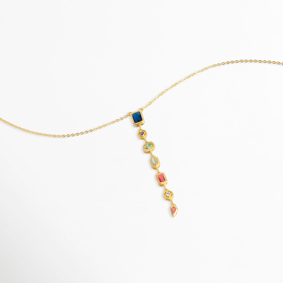 Toma en perspectiva del collar Prisma Dorado mostrando cómo cuelga el dije multicolor sobre la cadena dorada de 40 cm, Vista de lado del collar dorado con colgante vertical de piedras de color en caída suave, Estilismo del Collar Prisma Dorado en posición diagonal resaltando el cálido contraste del oro con las circonias multicolores, Muestra de la elegancia y movimiento del dije alargado multicolor enchapado en oro.