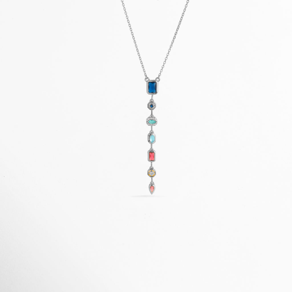 Collar Silver Prisma enchapado en oro blanco con dije colgante lineal vertical compuesto por siete circonias de colores en diferentes cortes, Vista de frente de collar plateado con colgante alargado estilo cascada de gemas multicolor sobre fondo blanco, Dije vertical de 7.5 cm con gemas de tonos azul, turquesa, fucsia, amarillo y rosa suspendido de una fina cadena plateada, Collar Silver Prisma con composición geométrica de circonias multicolores enmarcadas en biseles 