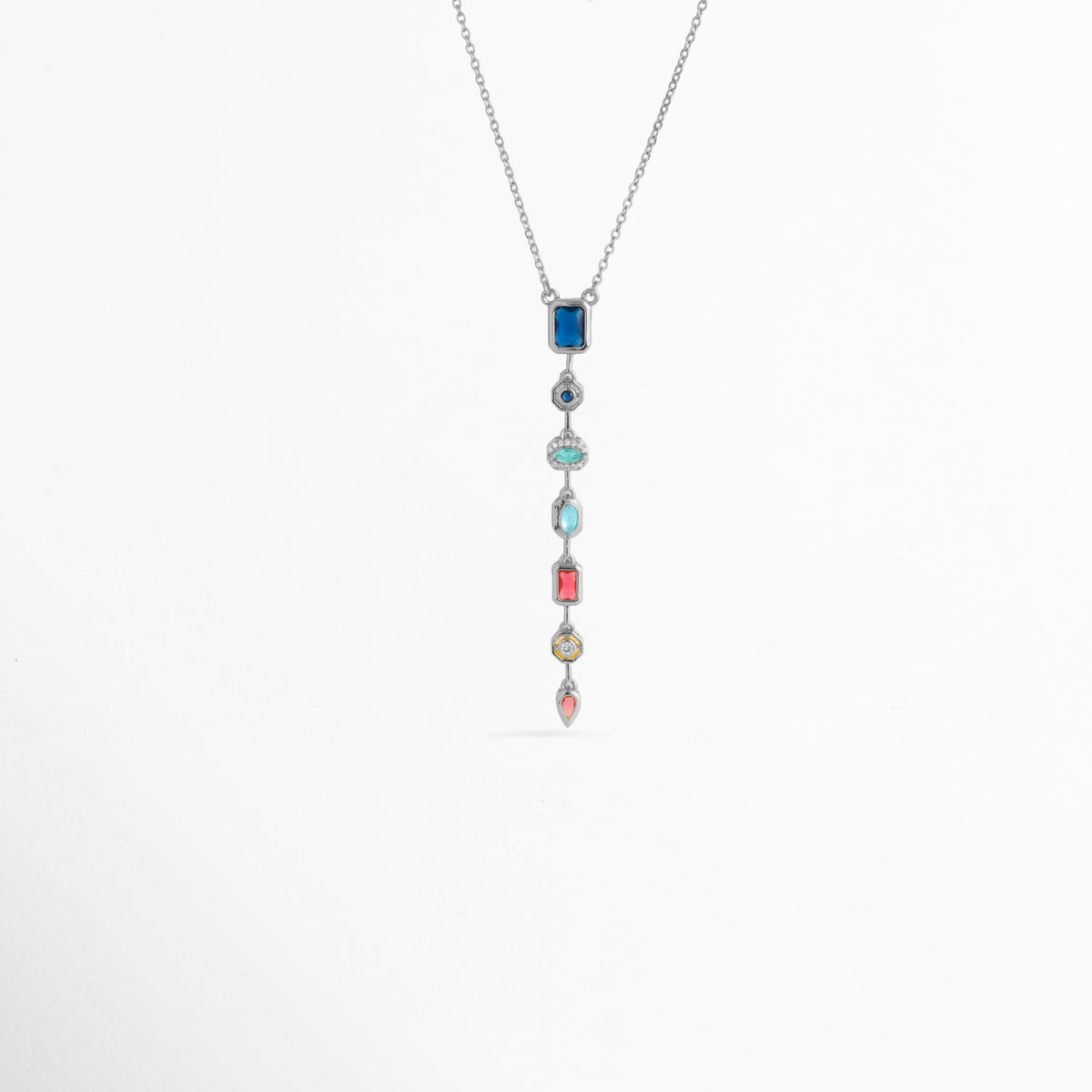 Collar Silver Prisma enchapado en oro blanco con dije colgante lineal vertical compuesto por siete circonias de colores en diferentes cortes, Vista de frente de collar plateado con colgante alargado estilo cascada de gemas multicolor sobre fondo blanco, Dije vertical de 7.5 cm con gemas de tonos azul, turquesa, fucsia, amarillo y rosa suspendido de una fina cadena plateada, Collar Silver Prisma con composición geométrica de circonias multicolores enmarcadas en biseles 