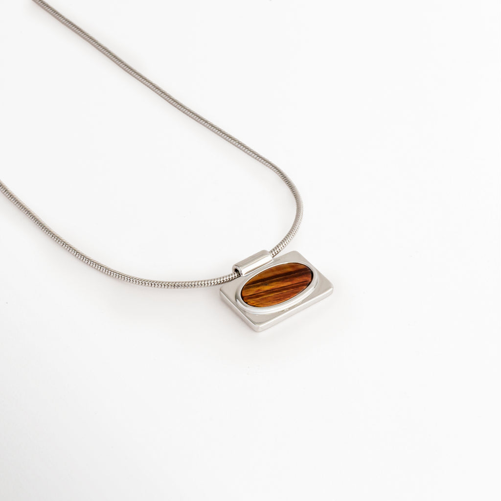 Vista en ángulo del Collar Tigra con colgante de piedra tipo Ojo de Tigre y cadena en acabado Oro Blanco, Perspectiva del Collar Tigra que resalta el brillo del colgante rectangular con piedra Ojo de Tigre, Collar Tigra fotografiado en ángulo mostrando el dije de piedra tipo Ojo de Tigre y su cadena fina, Vista diagonal del Collar Tigra con colgante elegante de piedra Ojo de Tigre, Collar Tigra con cadena de cobre y acabado en Oro Blanco visto desde una perspectiva lateral.
